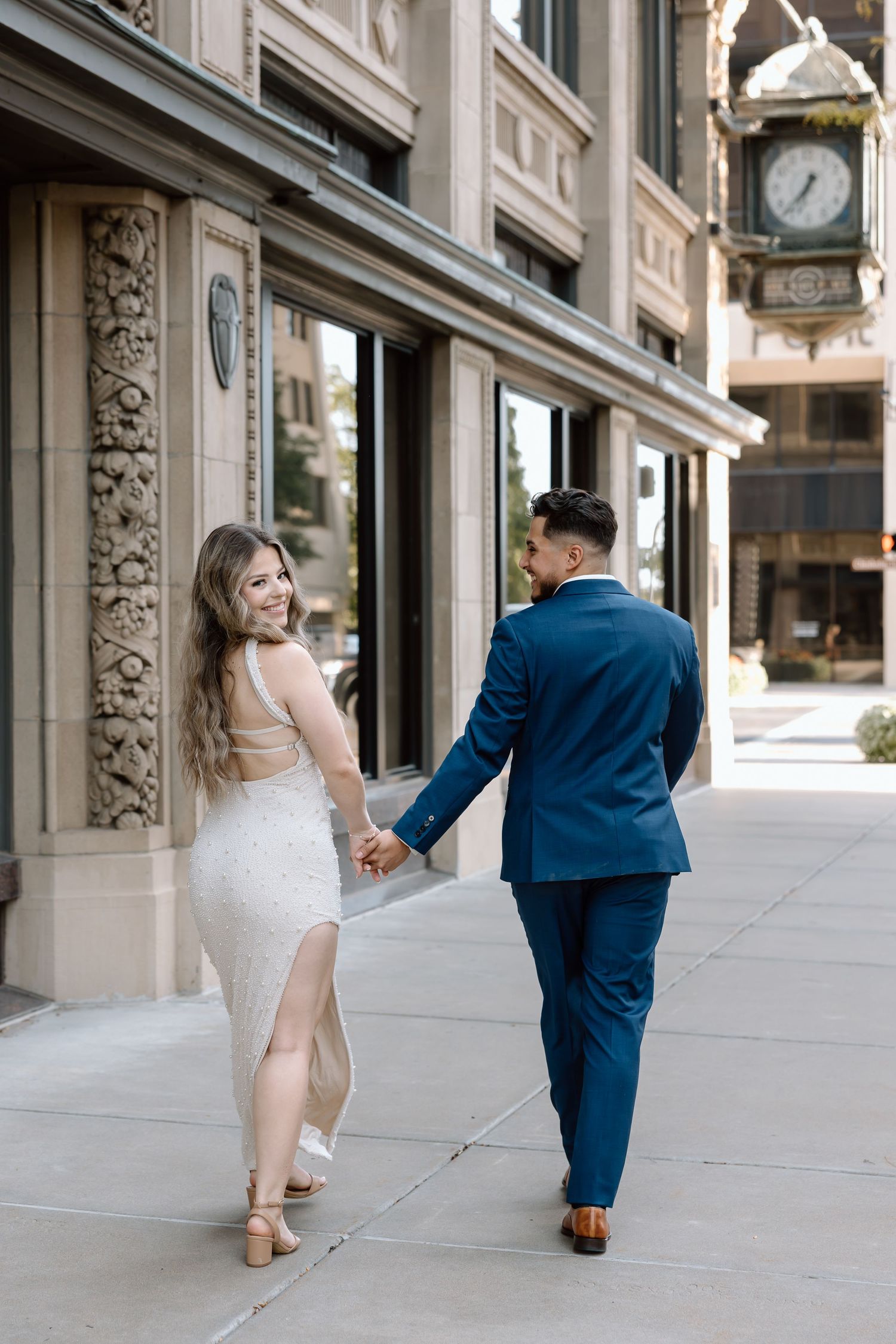Couple holding hands walking past ornate building with vintage clock downtown.