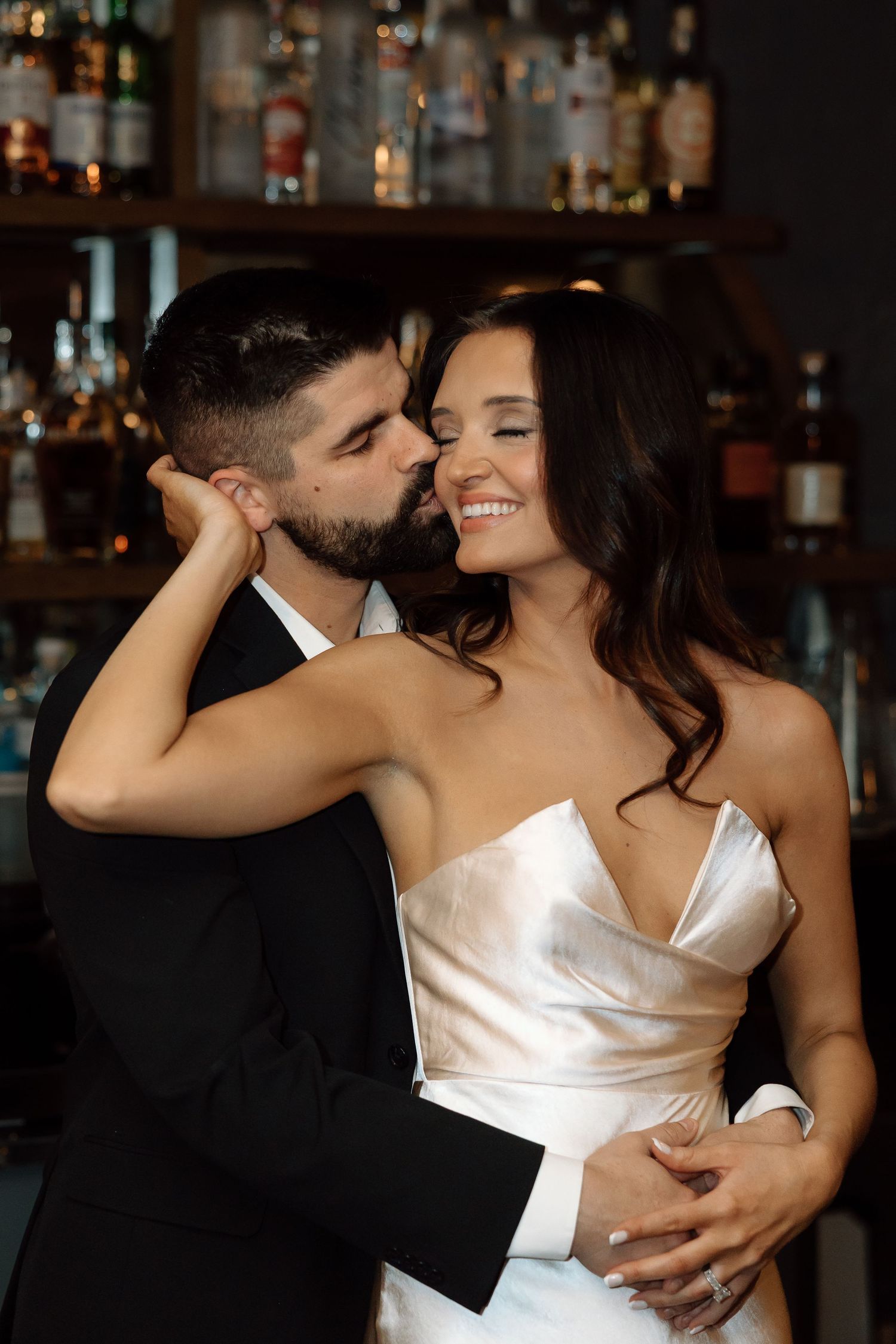 Groom in black suit kissing smiling bride in satin dress near a bar, romantic wedding portrait.