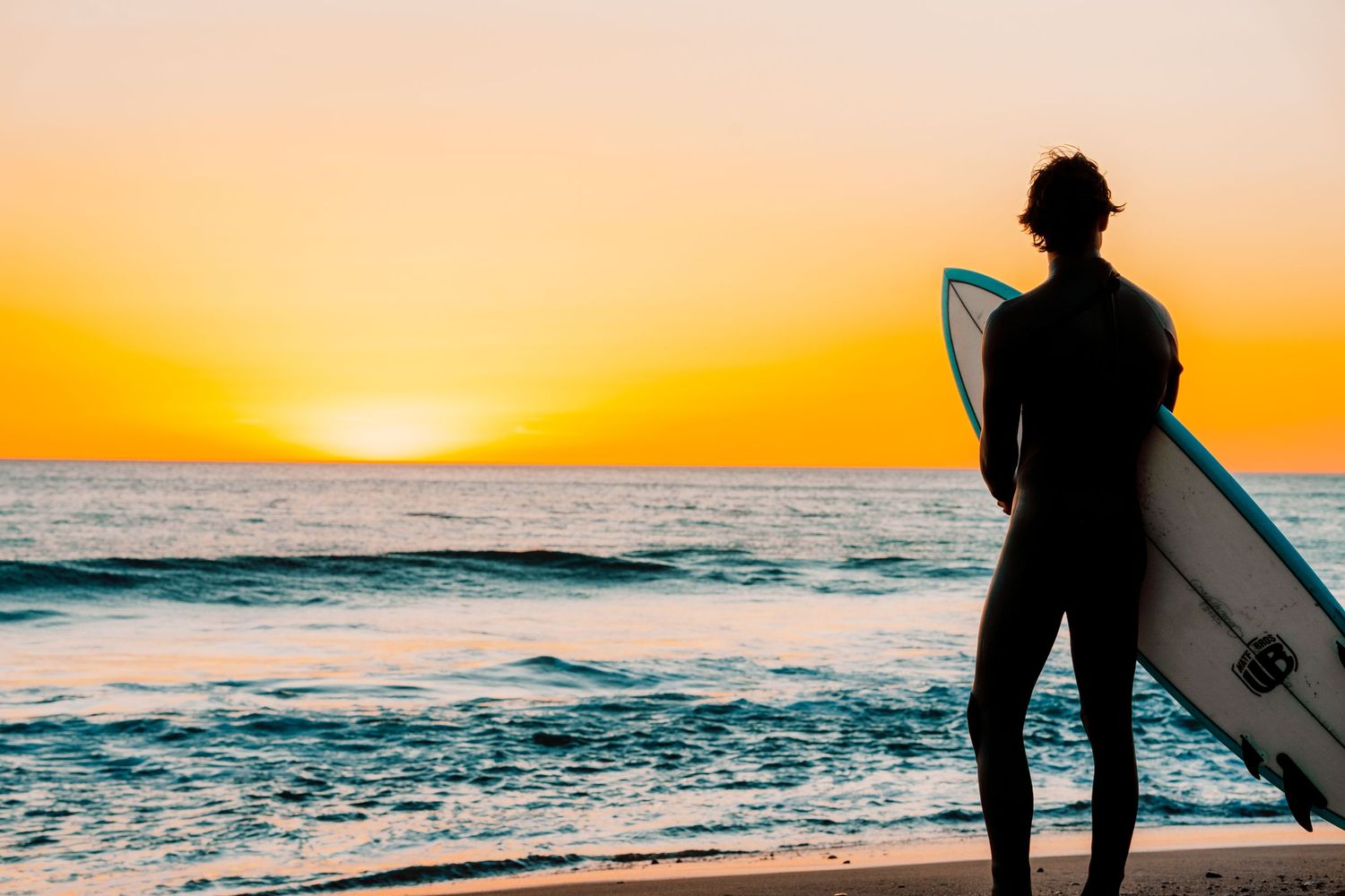 Silhouette of surfer with board watching golden sunset over ocean waves.