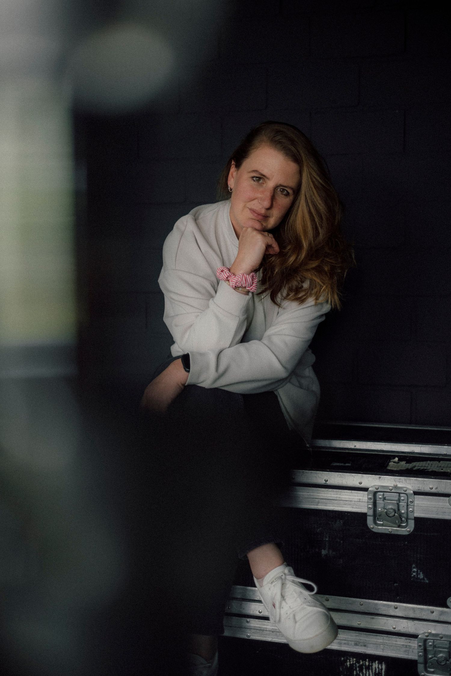 A person in a white sweater leans thoughtfully against a black piano in a moody, dark setting.