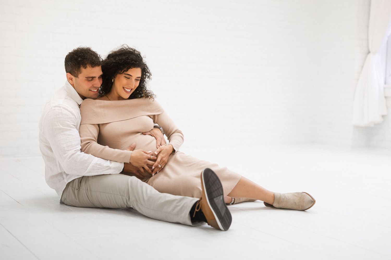 A couple sits together on a white floor, with the person in beige embracing their growing baby bump while resting peacefully.