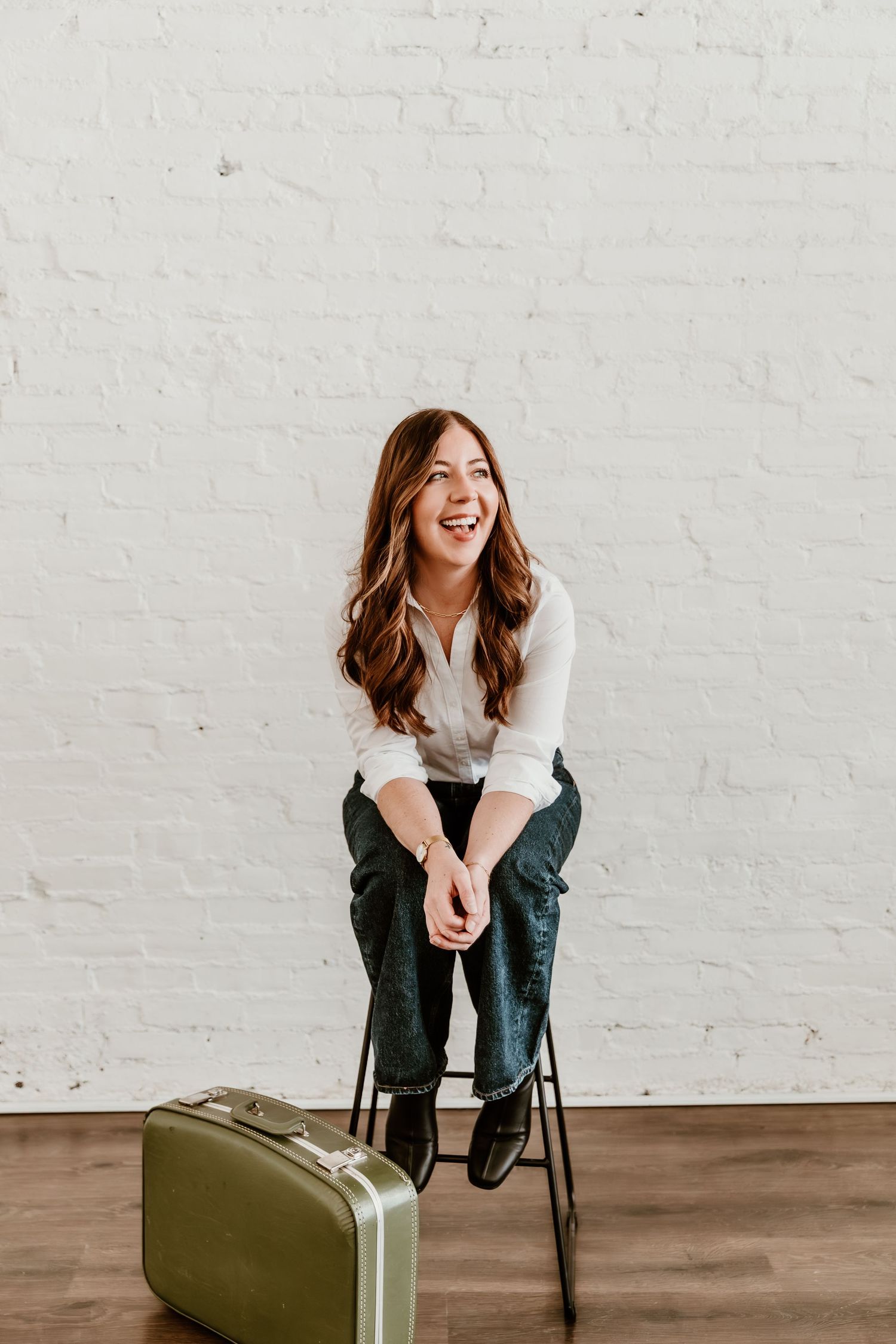 Woman in white shirt sitting on black chair laughing joyfully next to olive green vintage suitcase.