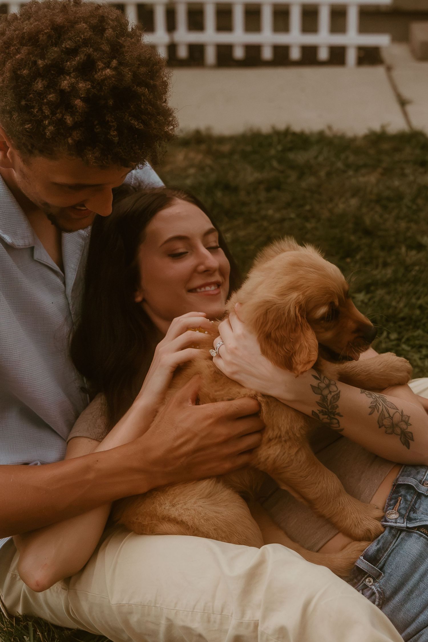A happy couple sits on grass cuddling a golden retriever puppy outdoors.
