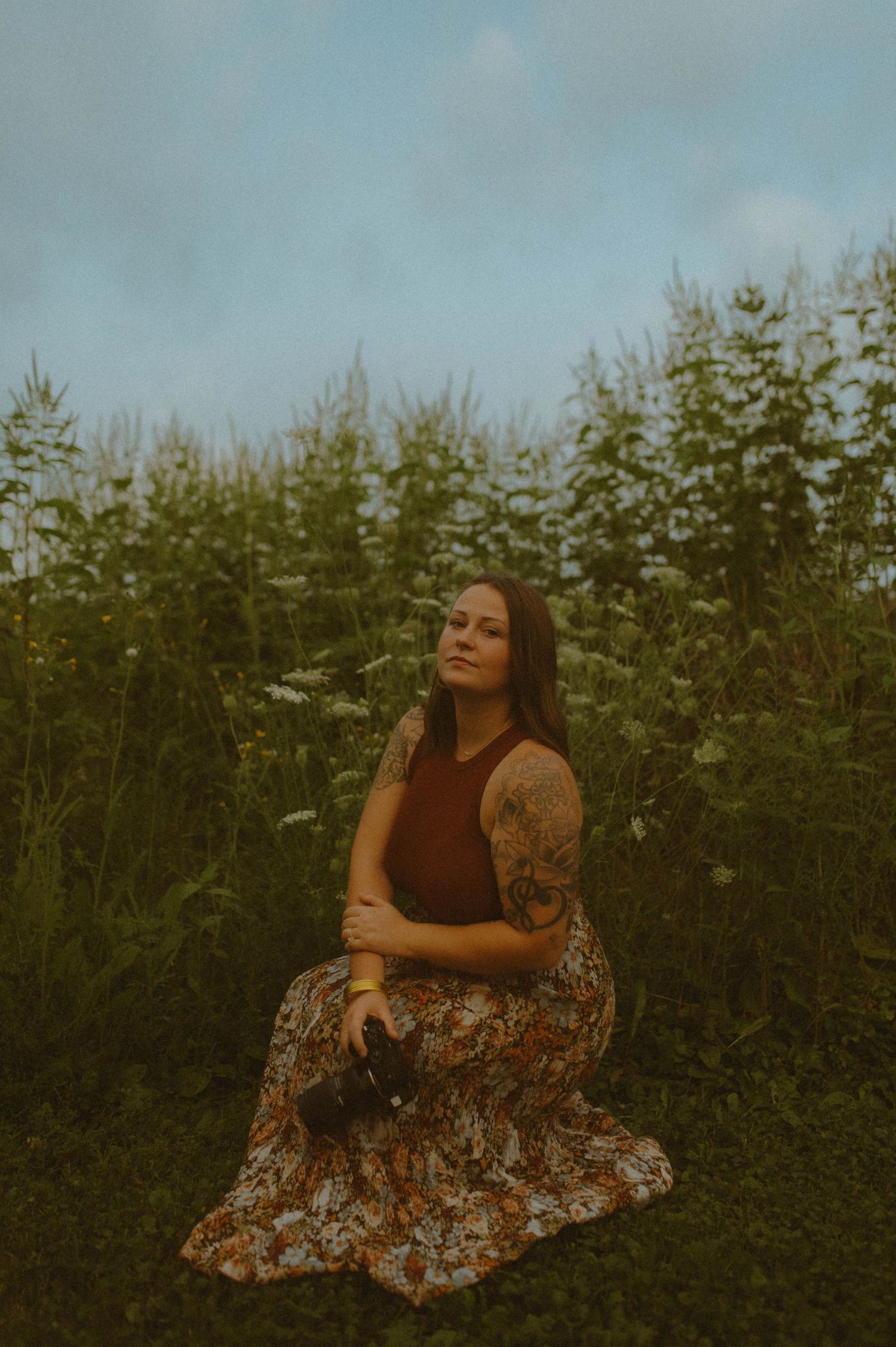 Tattooed woman in floral skirt sits peacefully in lush green field under cloudy sky.