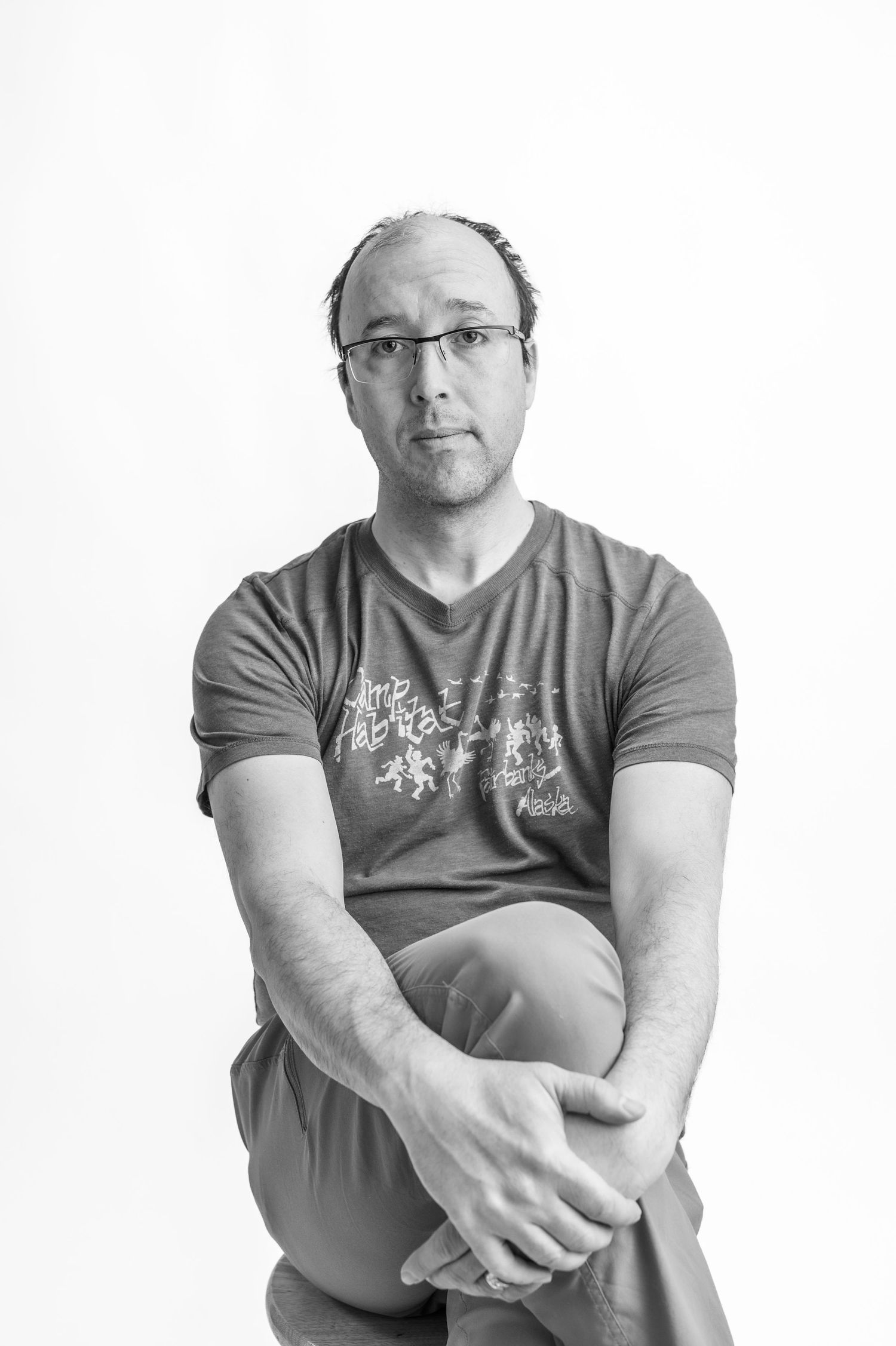 Black and white portrait of a man wearing glasses and a graphic t-shirt, sitting with hands clasped.