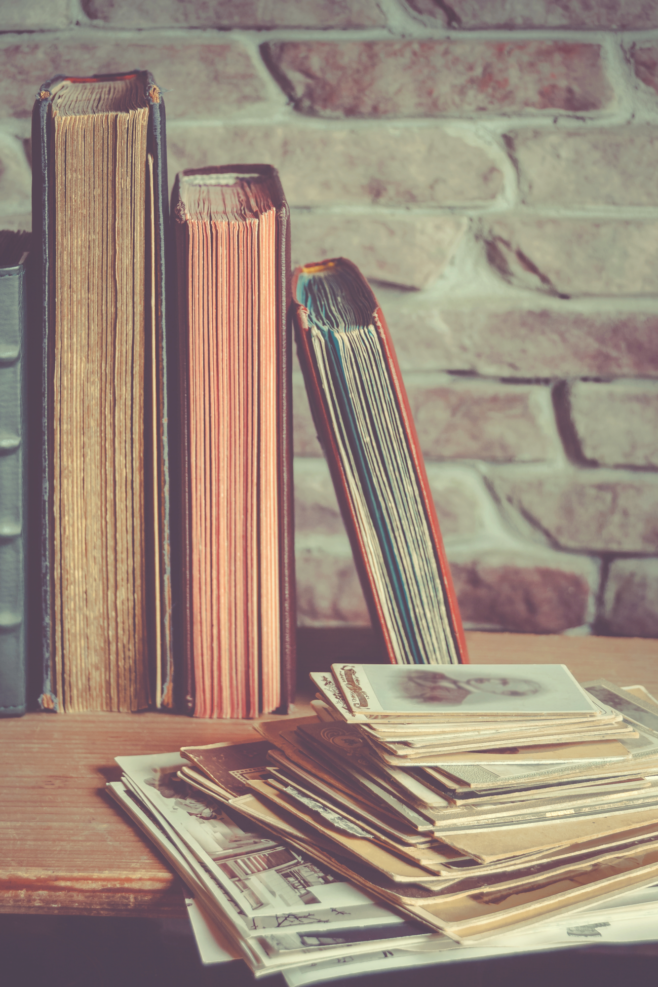 Stack of vintage photos leaning against old books on wooden table with brick wall in background.