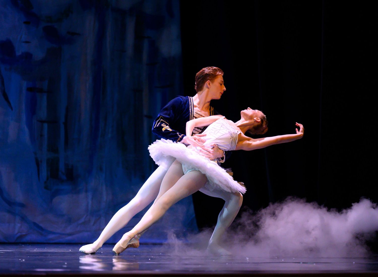 Ballet dancers perform graceful leaps and movements in white costumes on a misty stage during a performance.