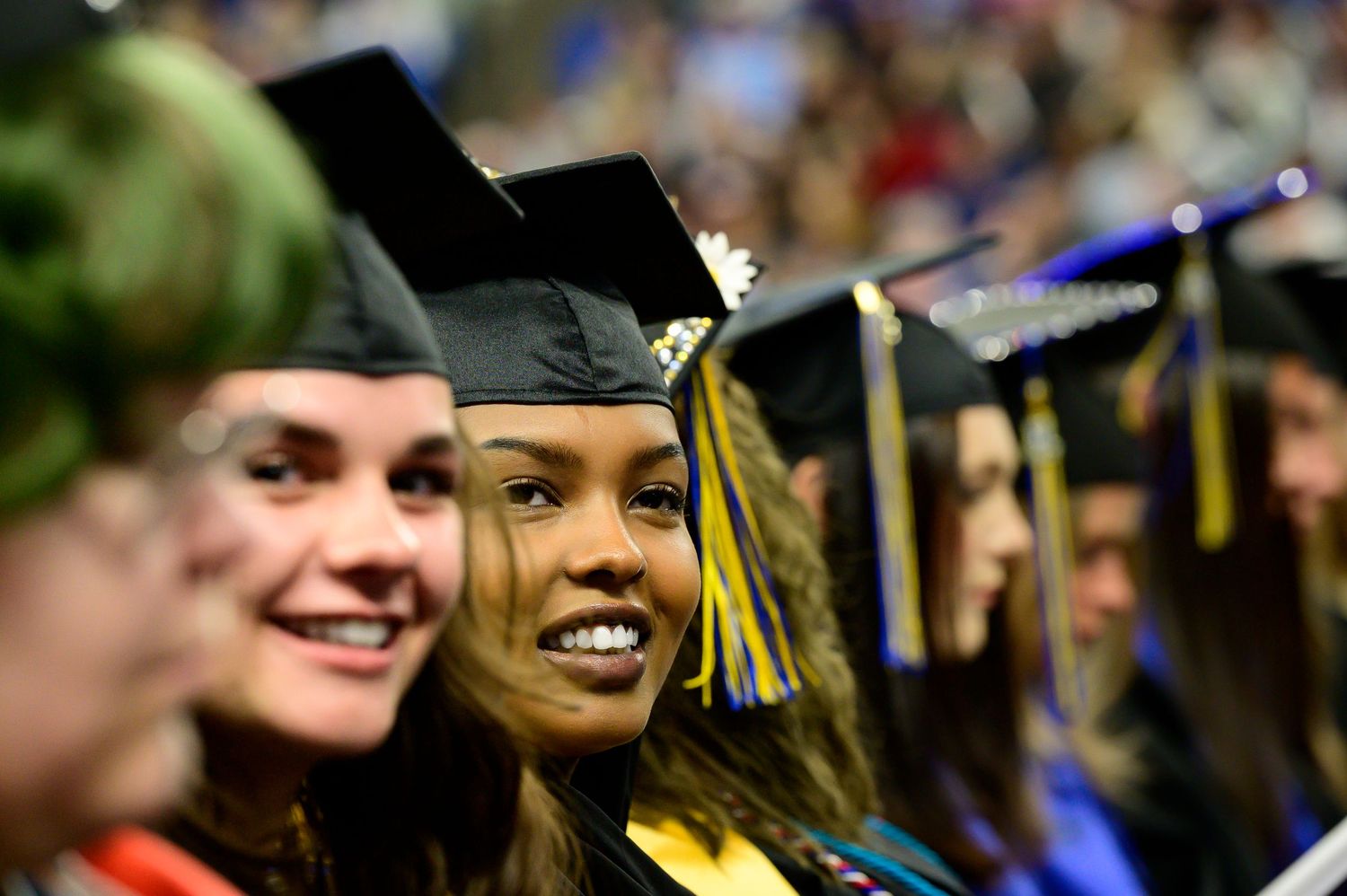 Graduates in black caps and gowns celebrate during a commencement ceremony with colorful lights and decorations.