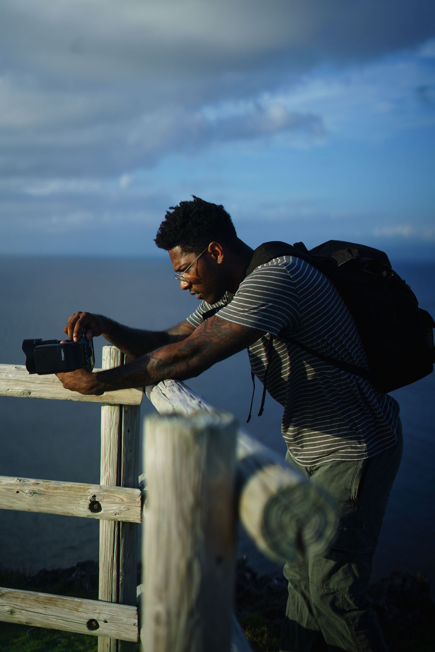 A photographer wearing a striped shirt and backpack takes photos from an oceanside viewing platform on a sunny day.
