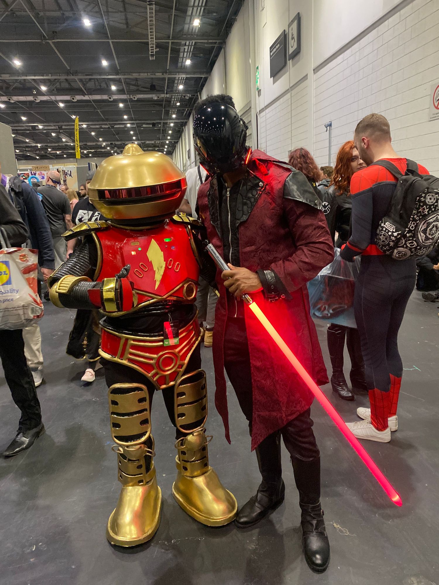 Two cosplayers dressed as sci-fi characters pose together at a convention, one in gold armor and one with a red lightsaber.