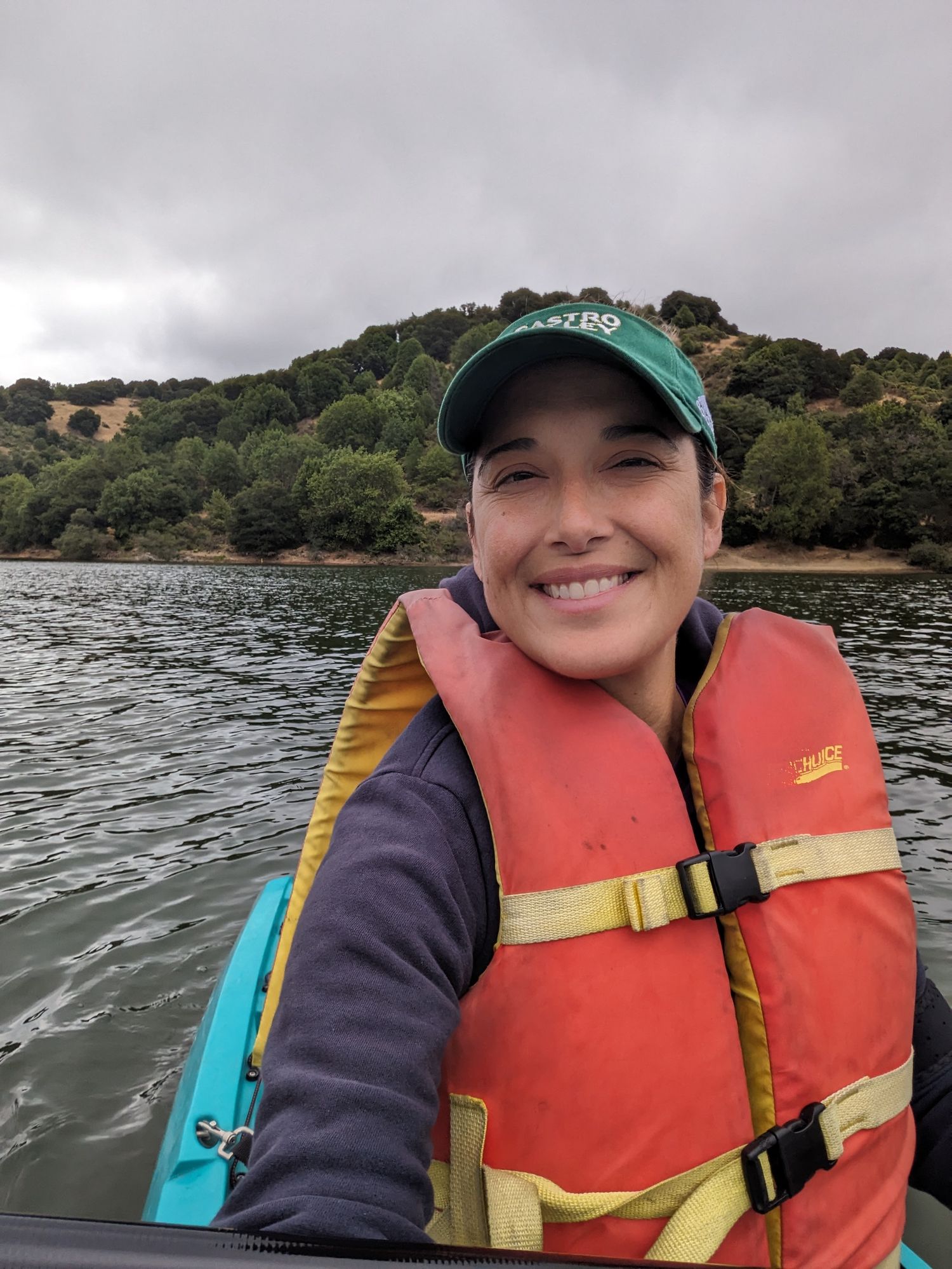 Someone wearing a green cap and orange life jacket enjoys kayaking on a lake under cloudy skies.