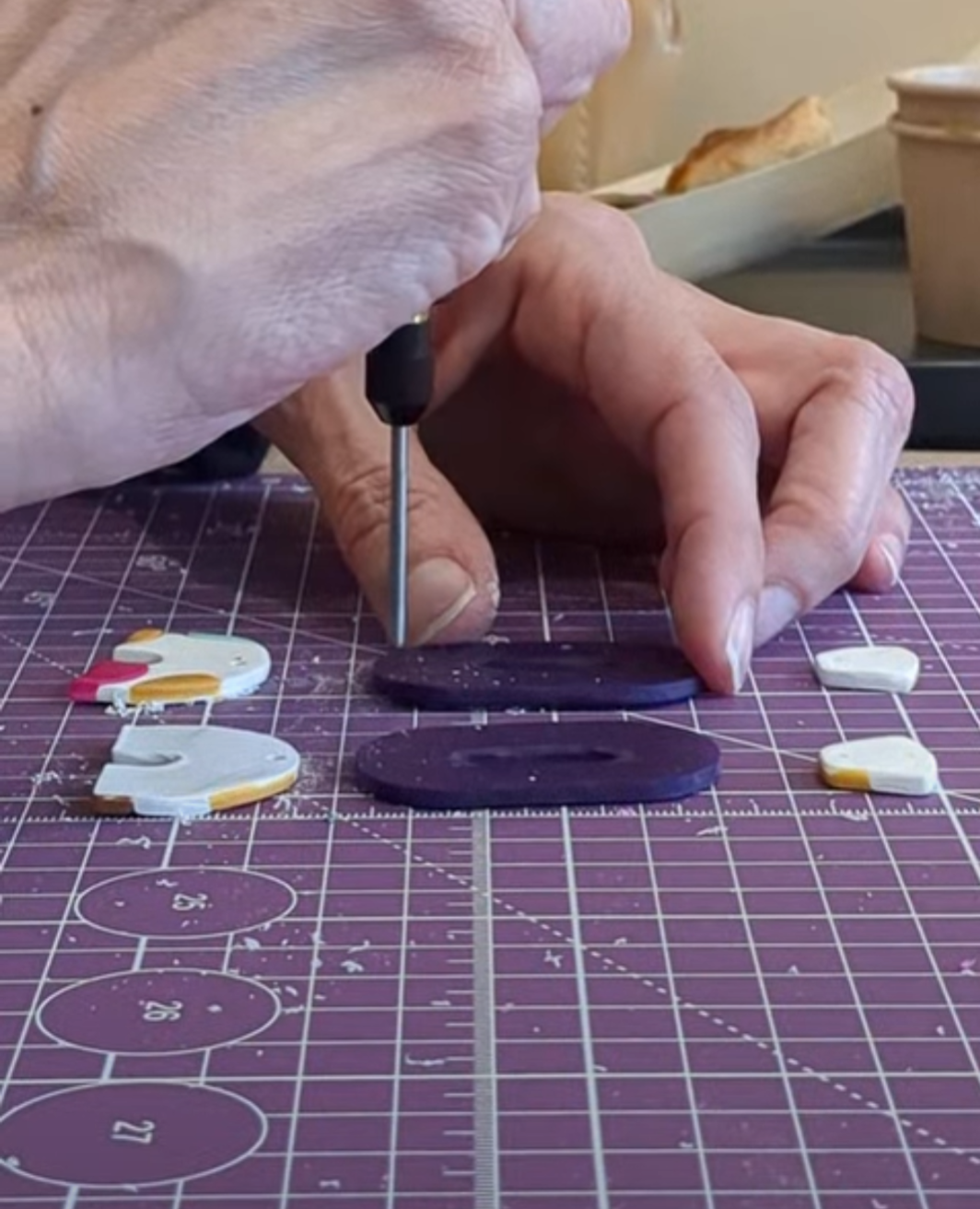 Hands working with craft tools on a purple cutting mat with small white stickers or elements visible.