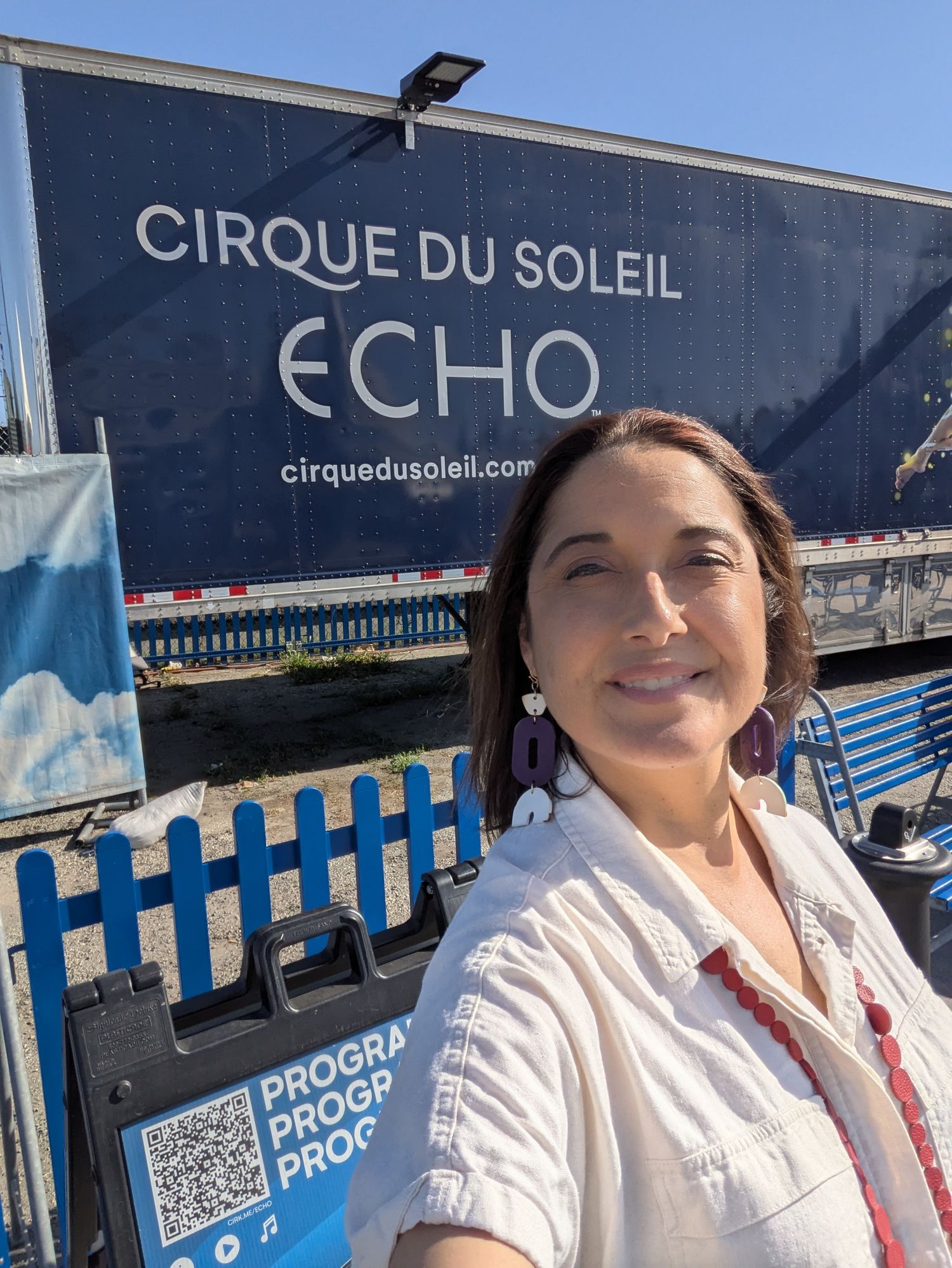 A person poses in front of a blue Cirque du Soleil Echo sign and blue fence with QR code display.