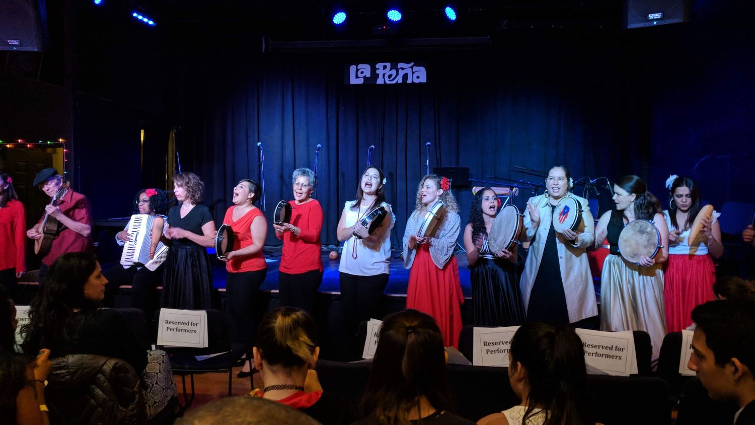 Students in red and black outfits perform together on stage at a musical recital under blue lighting.