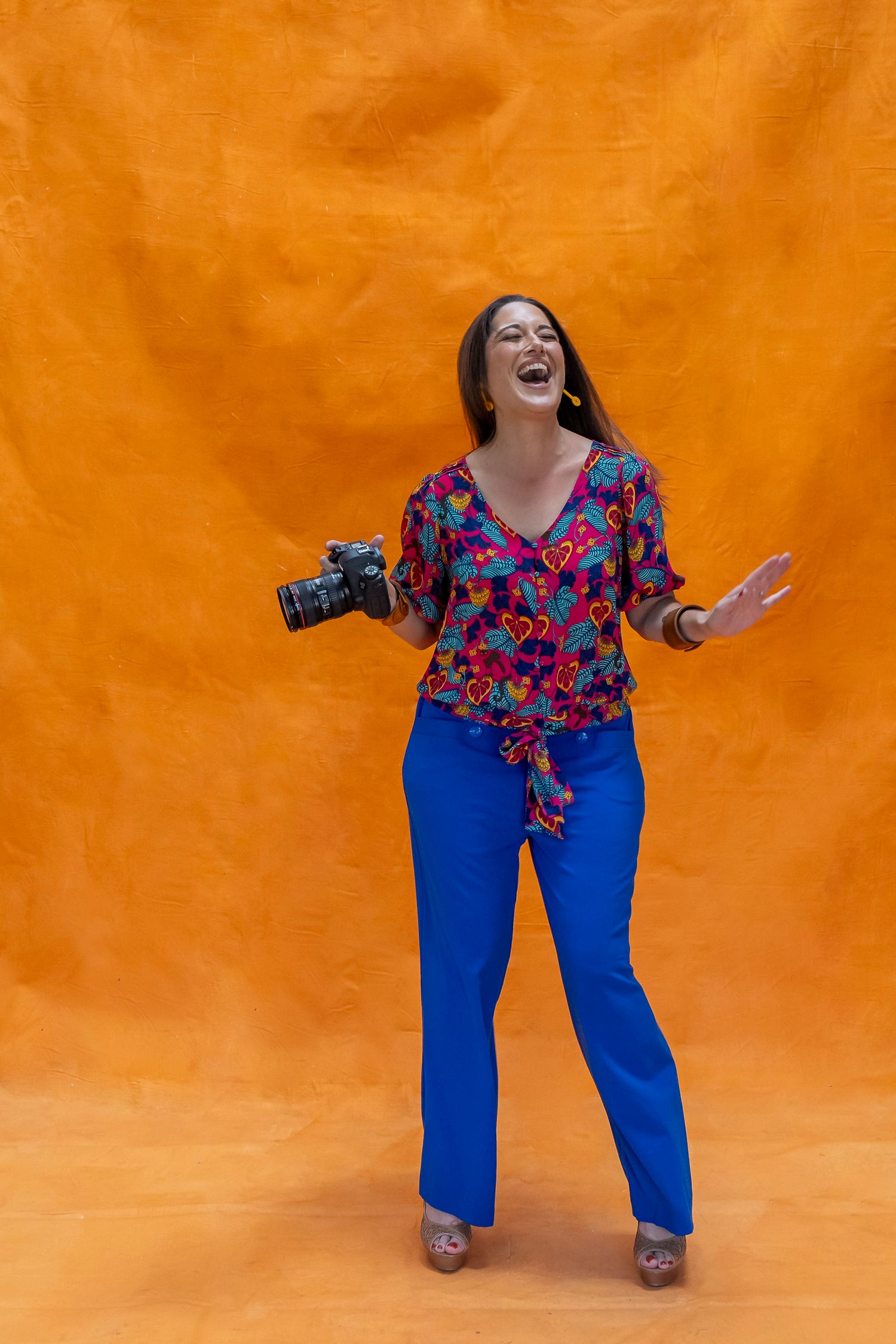 Photographer in a colorful floral top and blue pants poses joyfully against vibrant orange backdrop while holding camera.