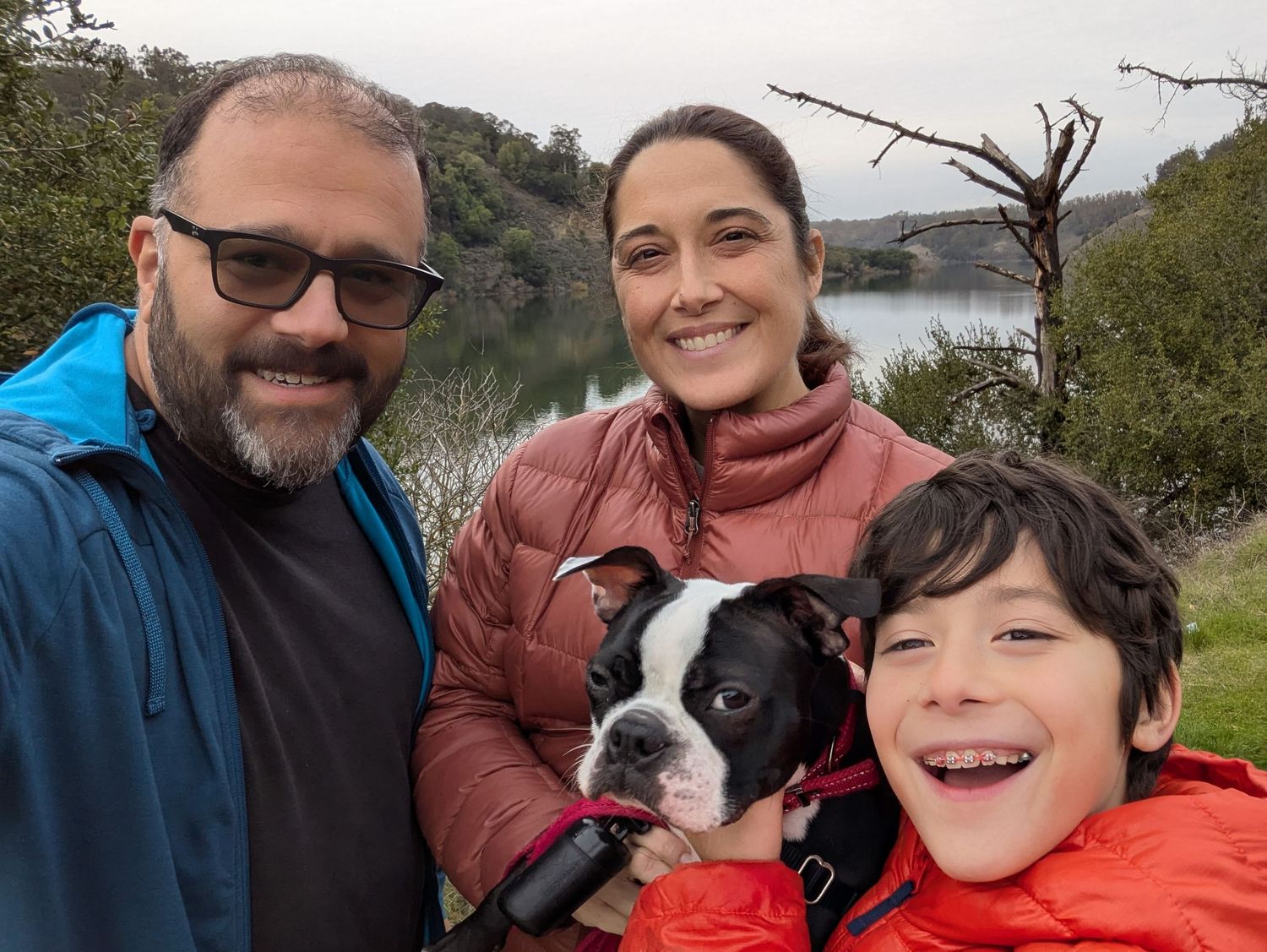 A family enjoying an outdoor hike near a lake with their Boston Terrier dog on a cloudy day.