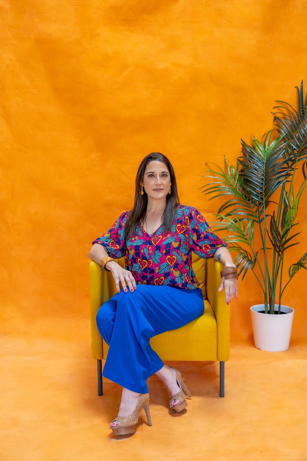 A person wearing a vibrant floral top and blue pants sits in a yellow chair against an orange wall with a potted palm.