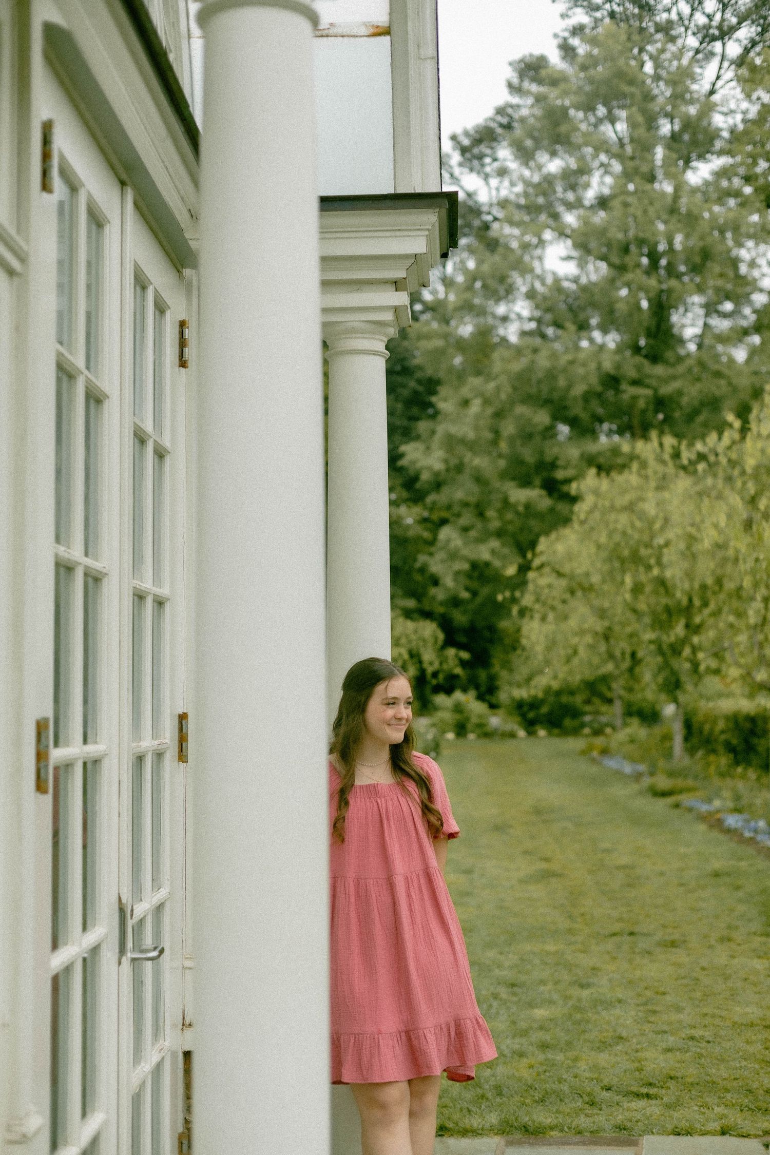 A coral dress flows elegantly against white colonial columns and green landscaping.