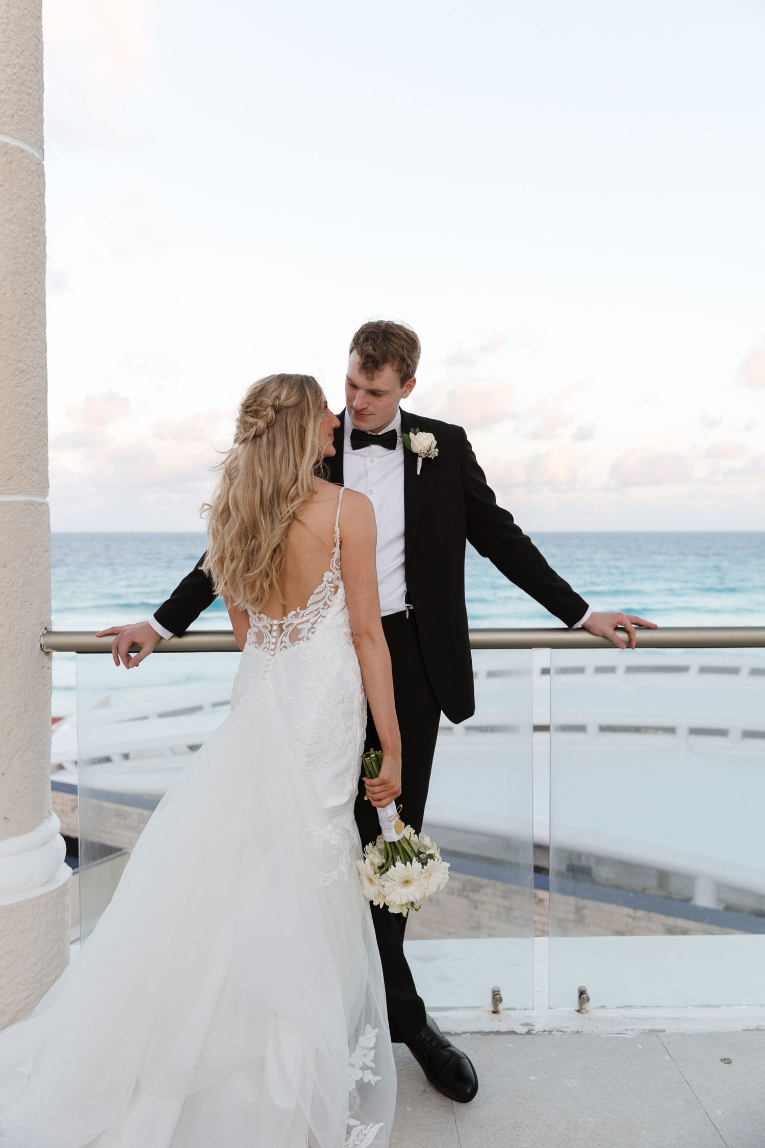 Bride and groom share a romantic moment on an oceanfront balcony, she in a lace gown holding white flowers, he in a black tuxedo.