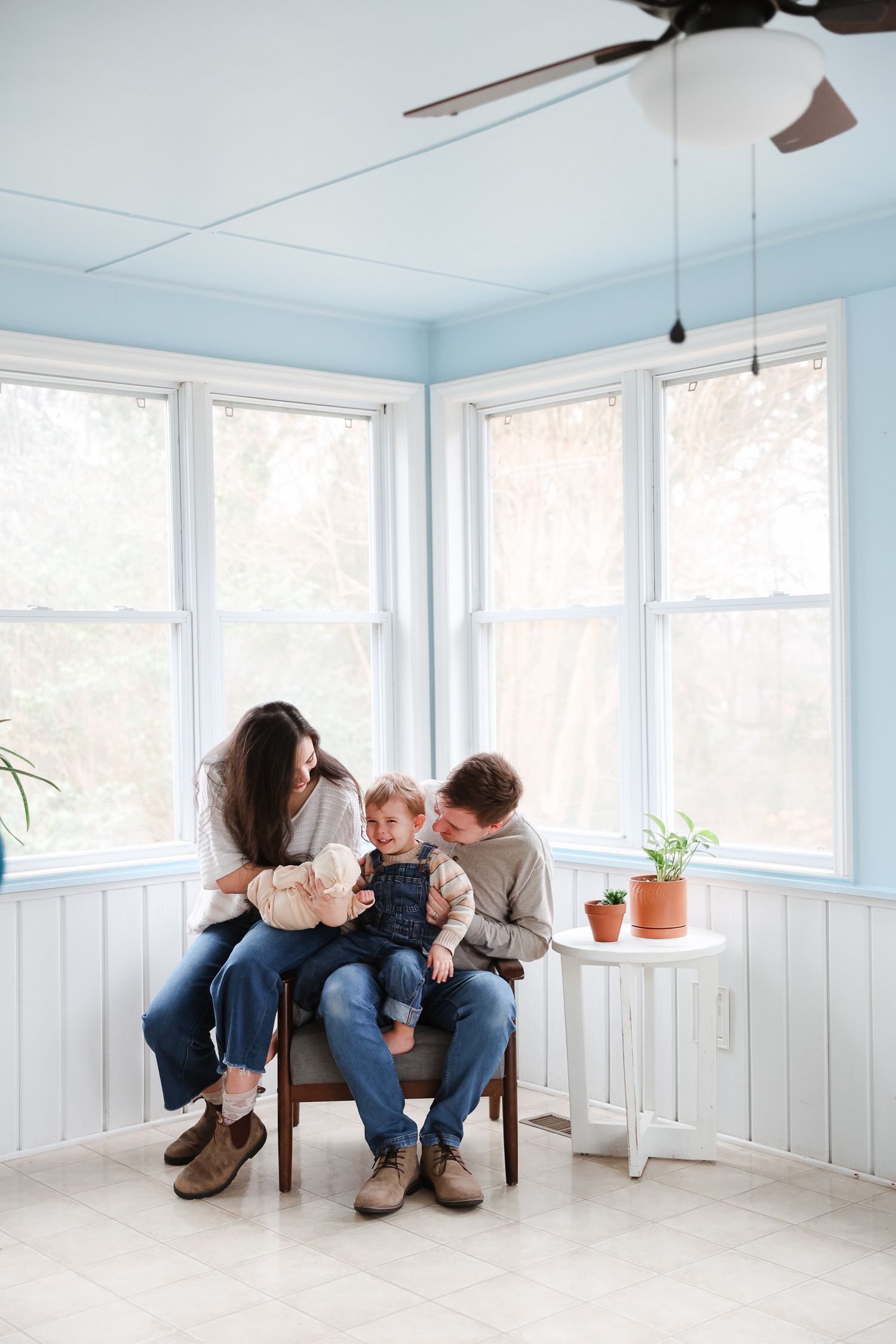 Family of three sitting together on a chair in a bright sunroom with blue ceiling and white wainscoting.