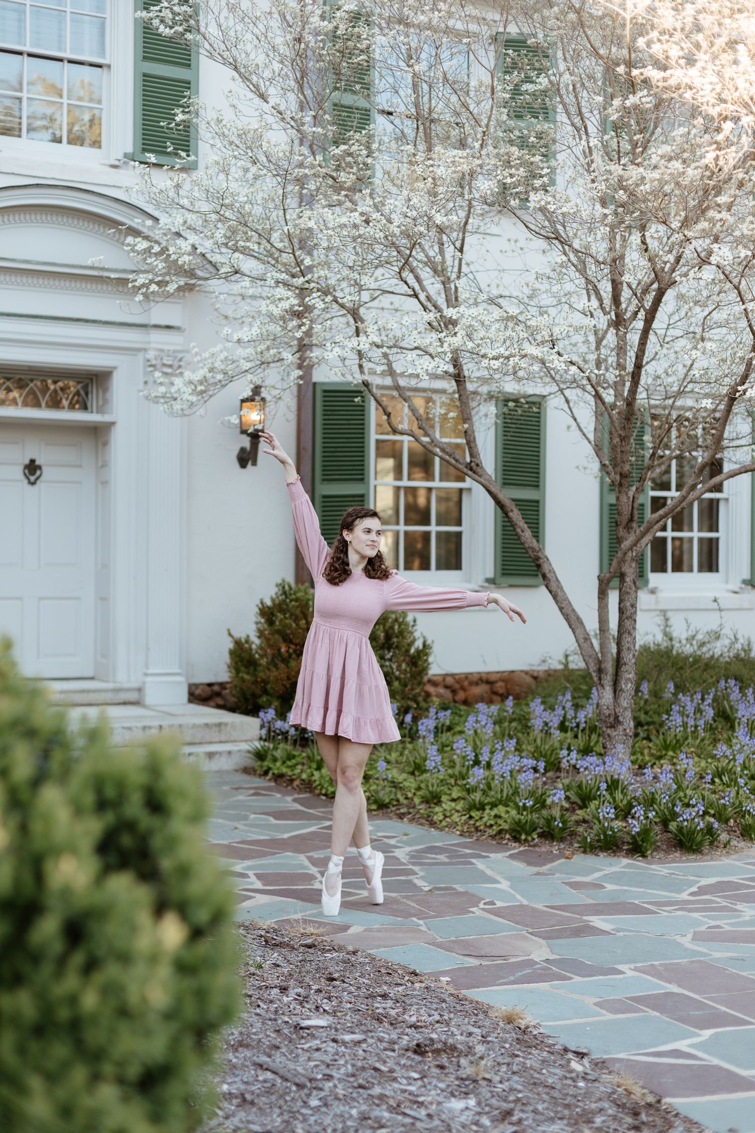 A flowing pink dress moves gracefully against a white building with blooming spring trees.