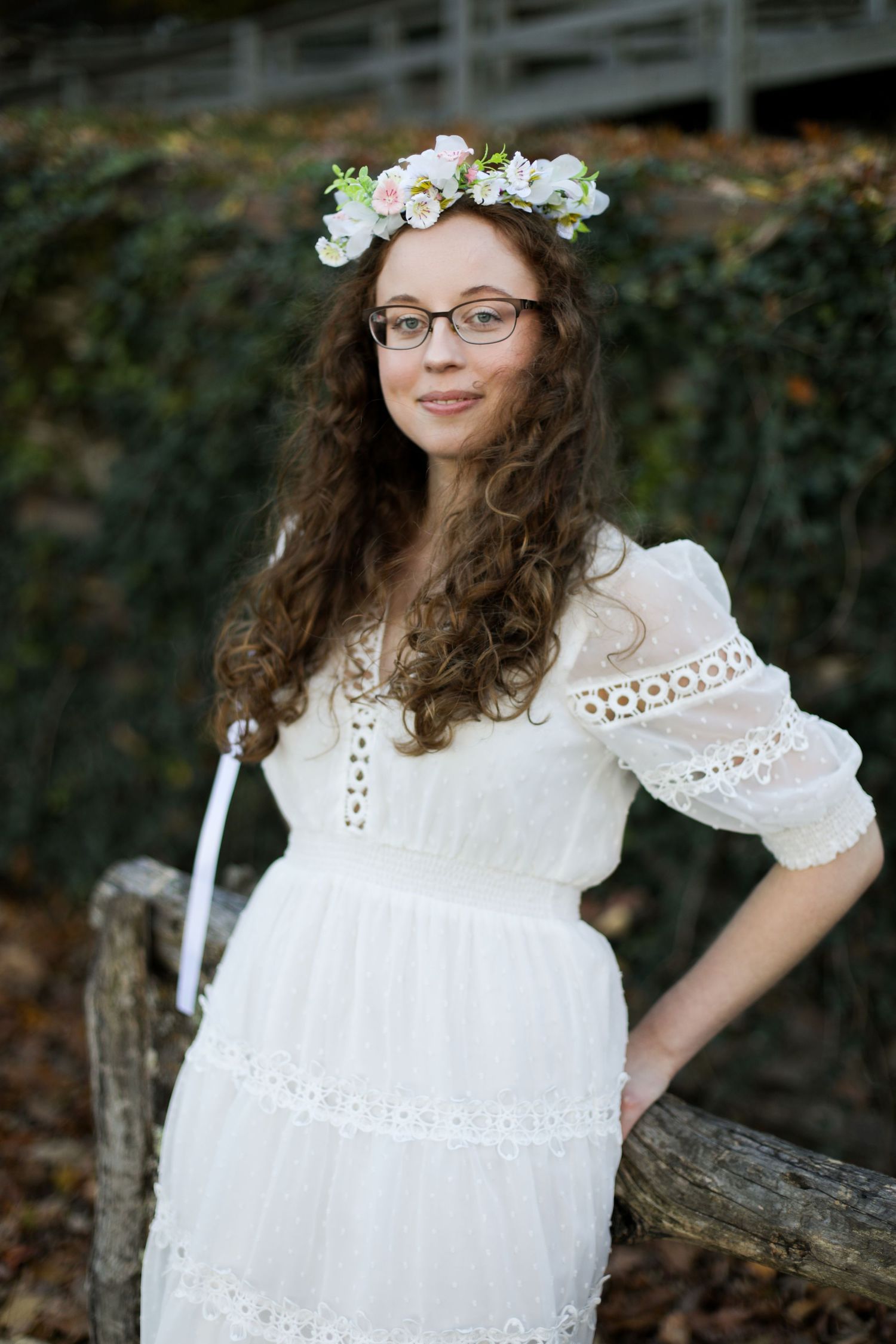 Young person in white eyelet dress and floral crown poses outdoors.