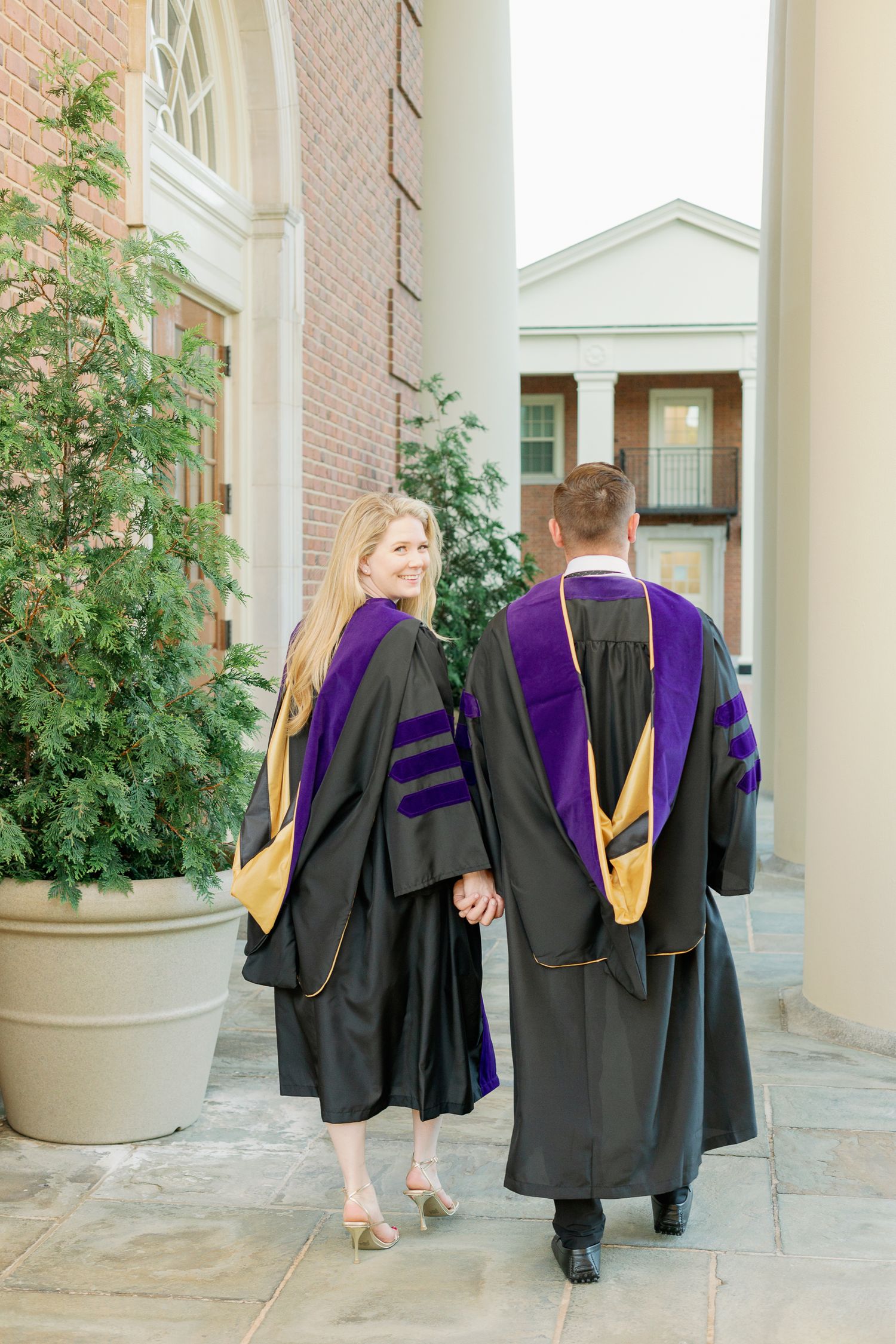 Graduates in purple and black academic regalia walking and posing between architectural columns.