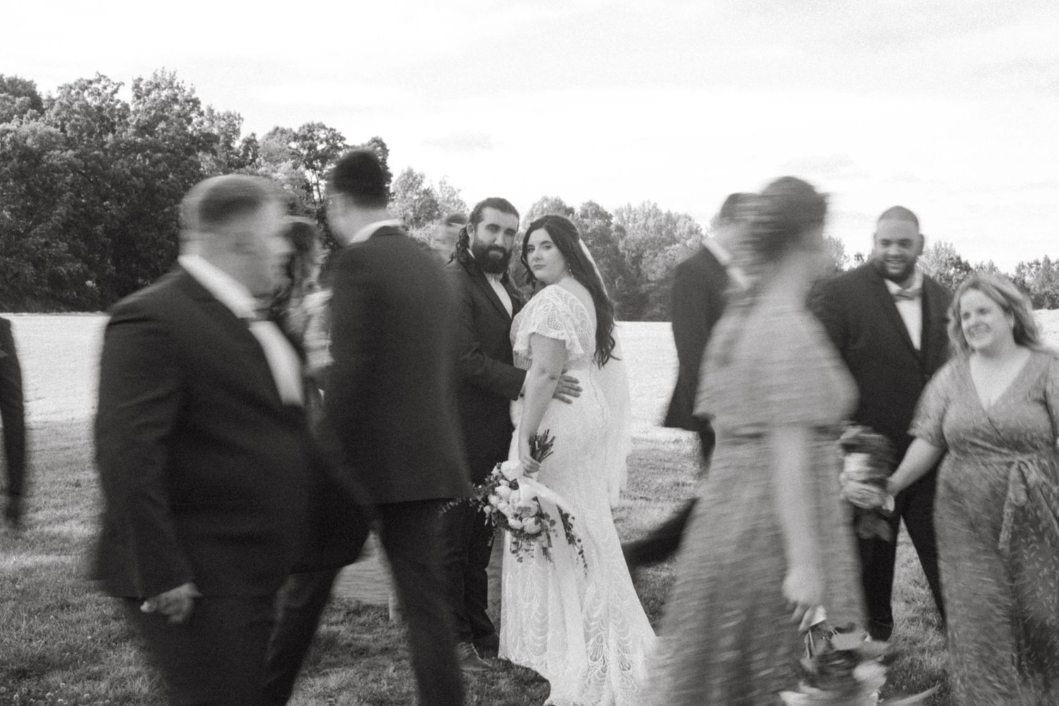 A black and white photo of wedding guests mingling outdoors on a sunny day.