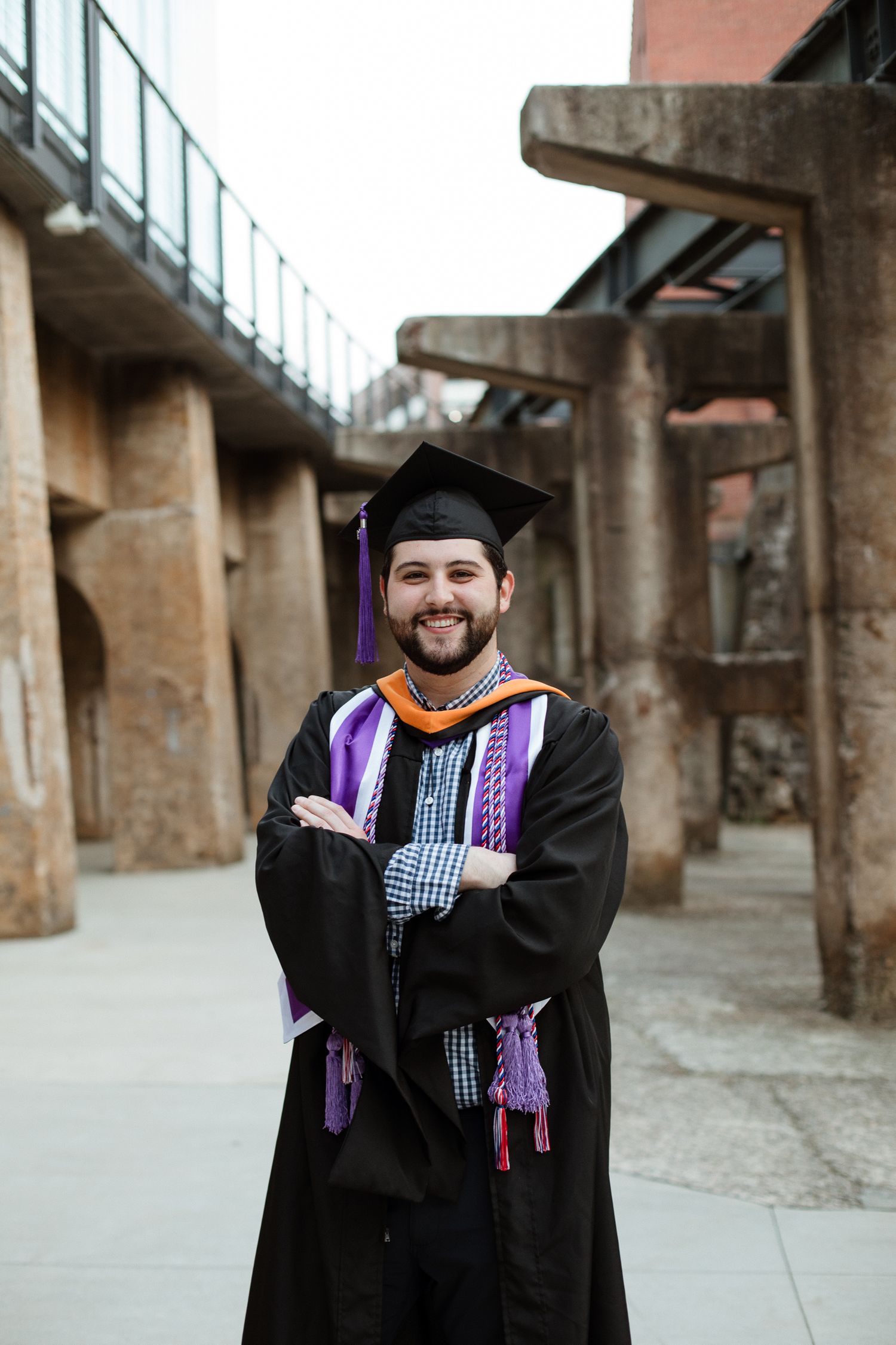 A graduate in cap and gown with purple stole poses in front of industrial architecture.
