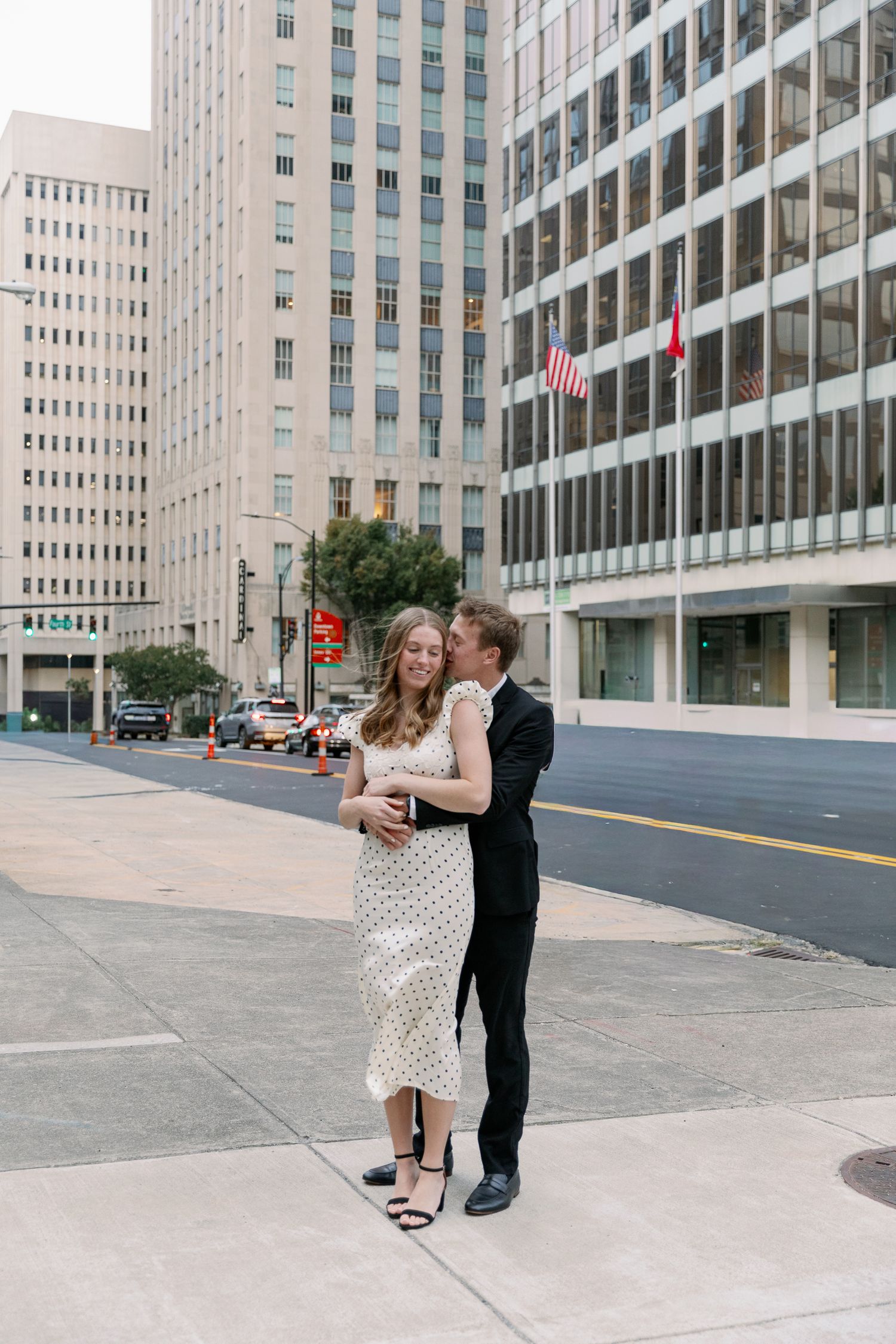 A couple embraces on a city sidewalk with modern office buildings and an American flag in the background.