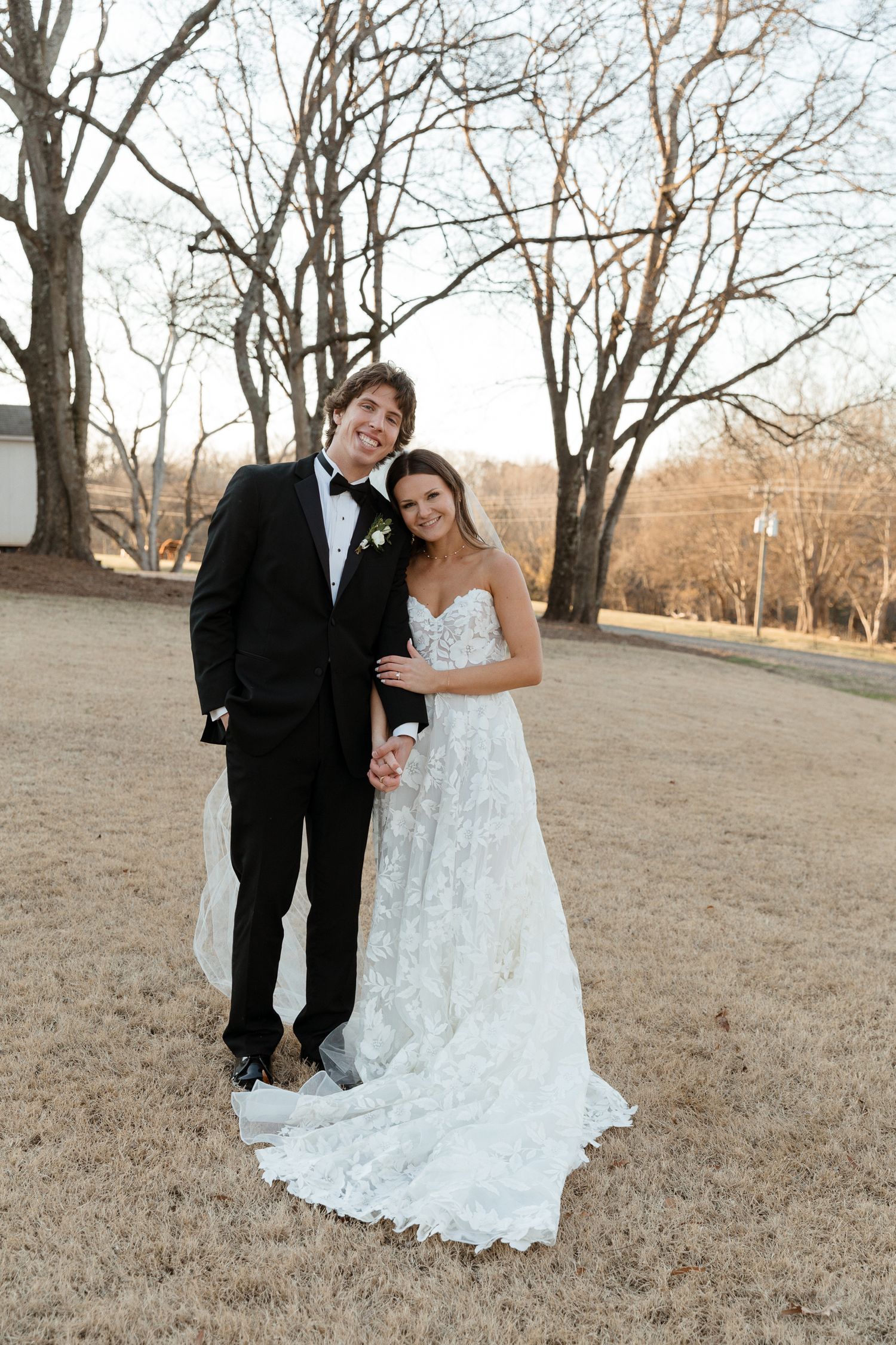 A couple poses together in a winter field with bare trees in the background.