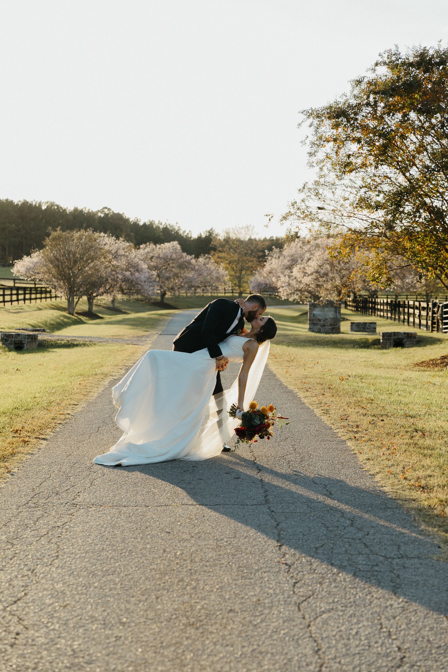 A couple shares a romantic moment on a path surrounded by blooming cherry trees and wooden fences at sunset.