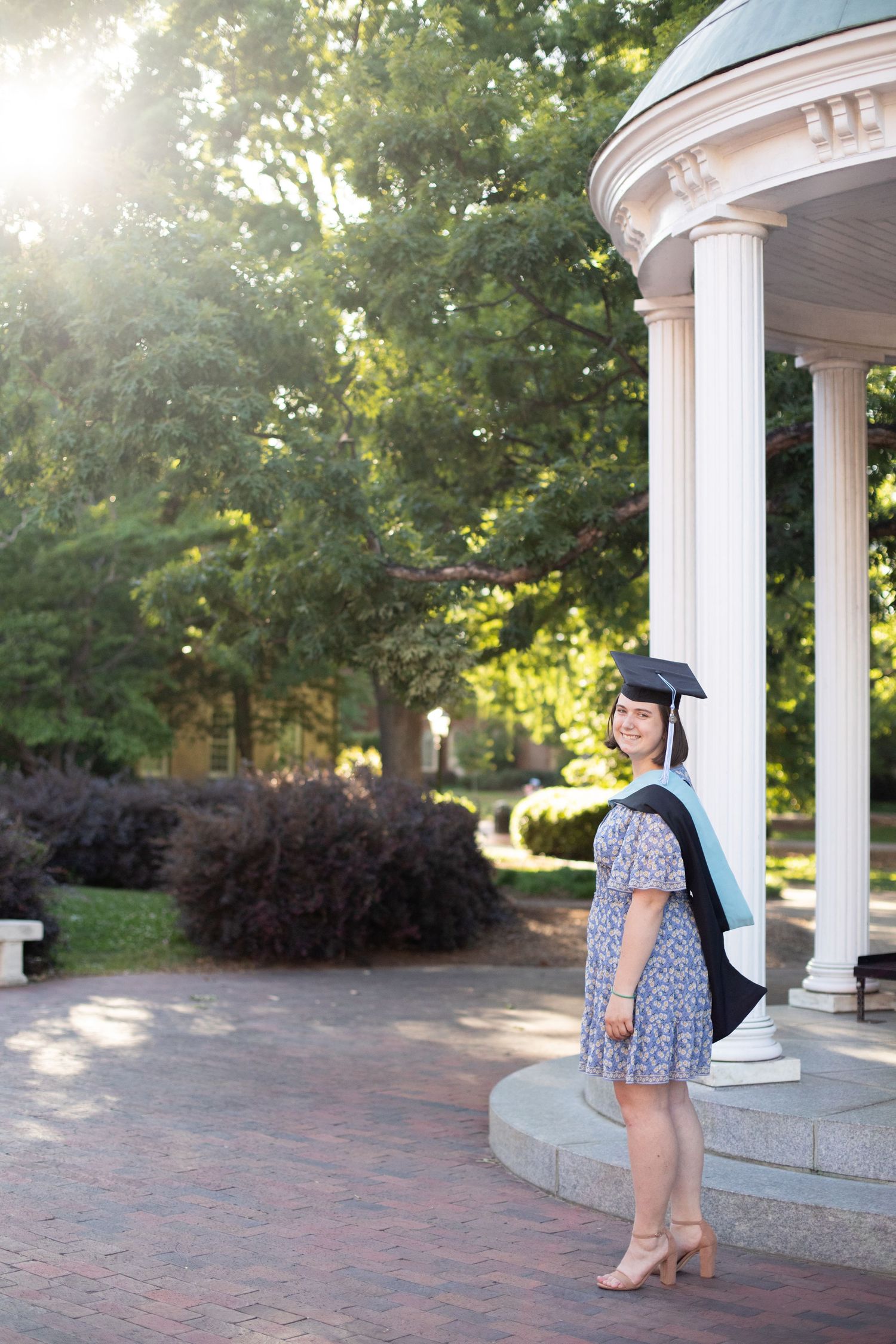 A graduate poses in cap and gown by white classical columns during golden hour.
