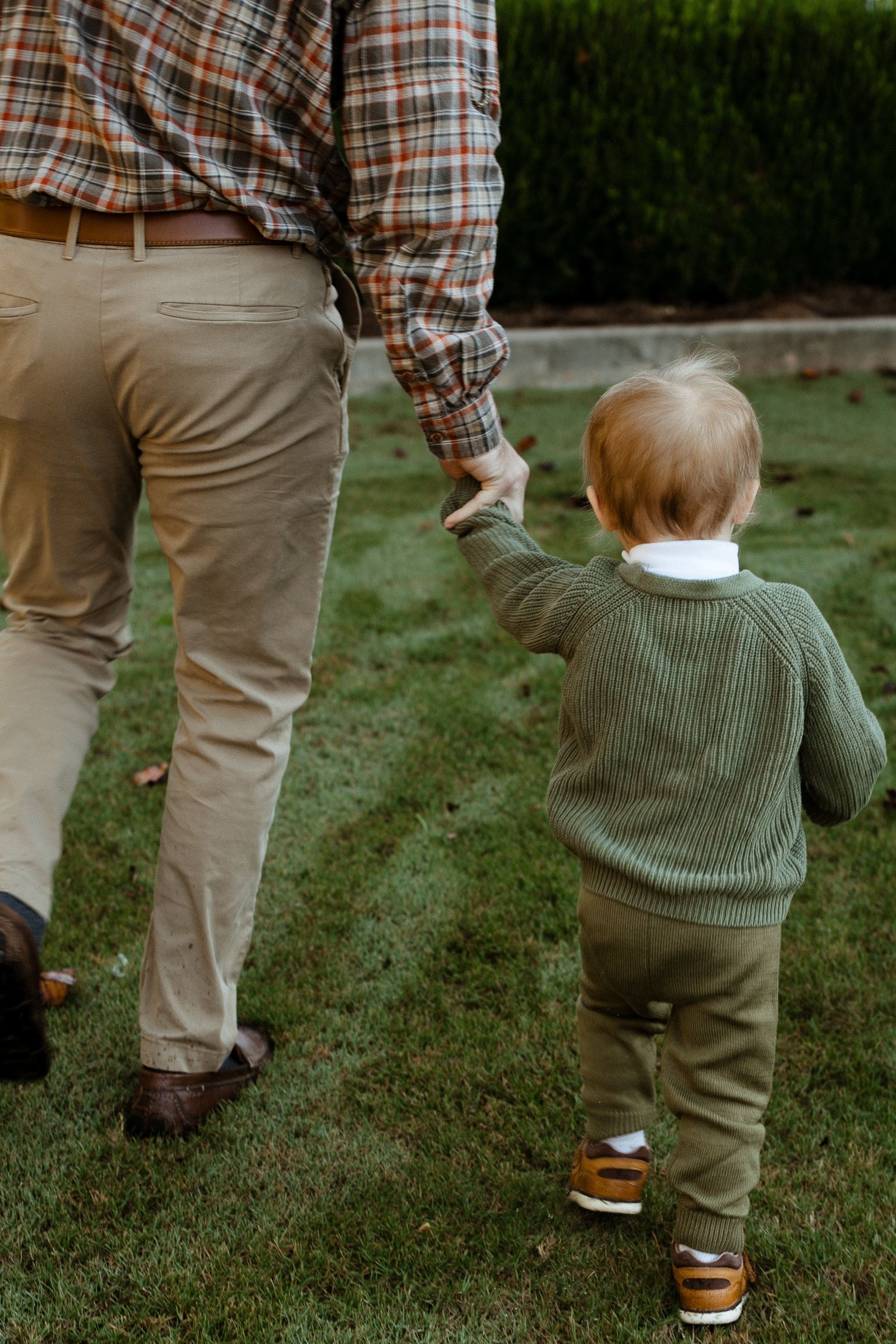 A small child in a green sweater follows behind an adult wearing plaid and khakis on grassy ground.