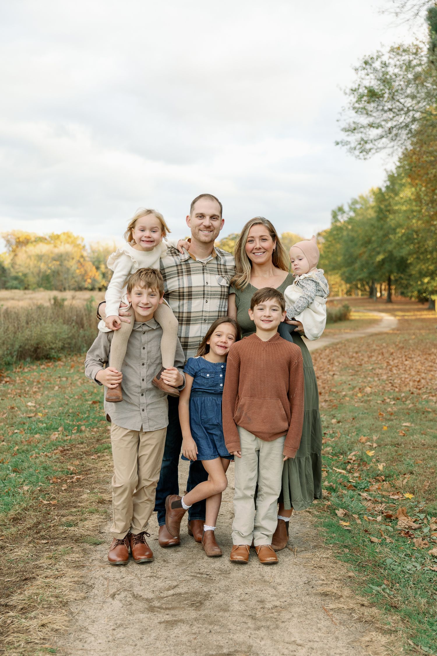 Family of six poses on a scenic autumn trail, parents with four children in coordinated fall outfits, surrounded by colorful foliage.