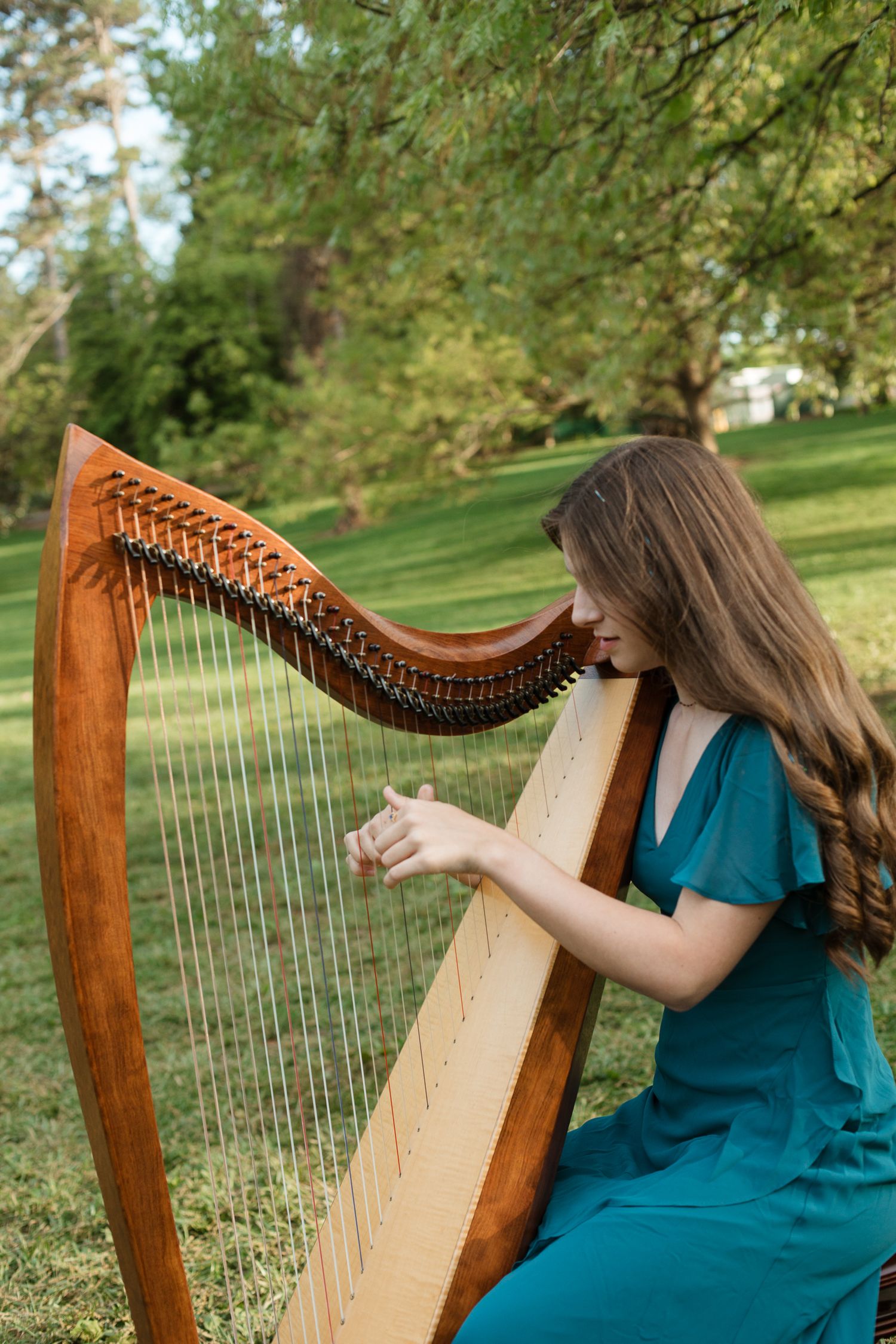 A harpist in a teal dress performs outdoors in a lush green garden setting.