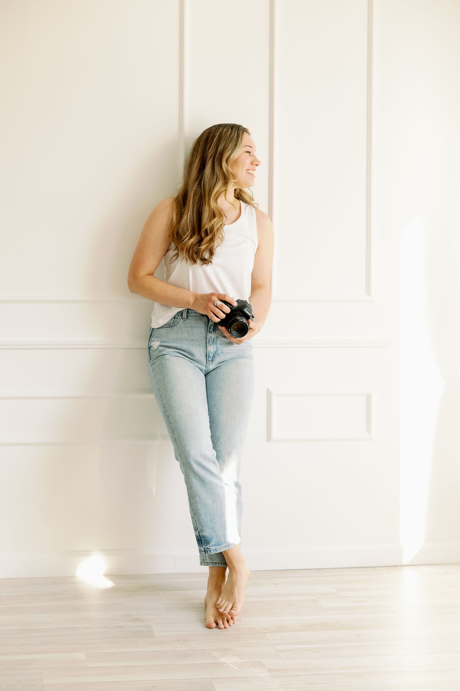 Relaxed barefoot pose against white wall wearing classic denim and white tank while holding camera.