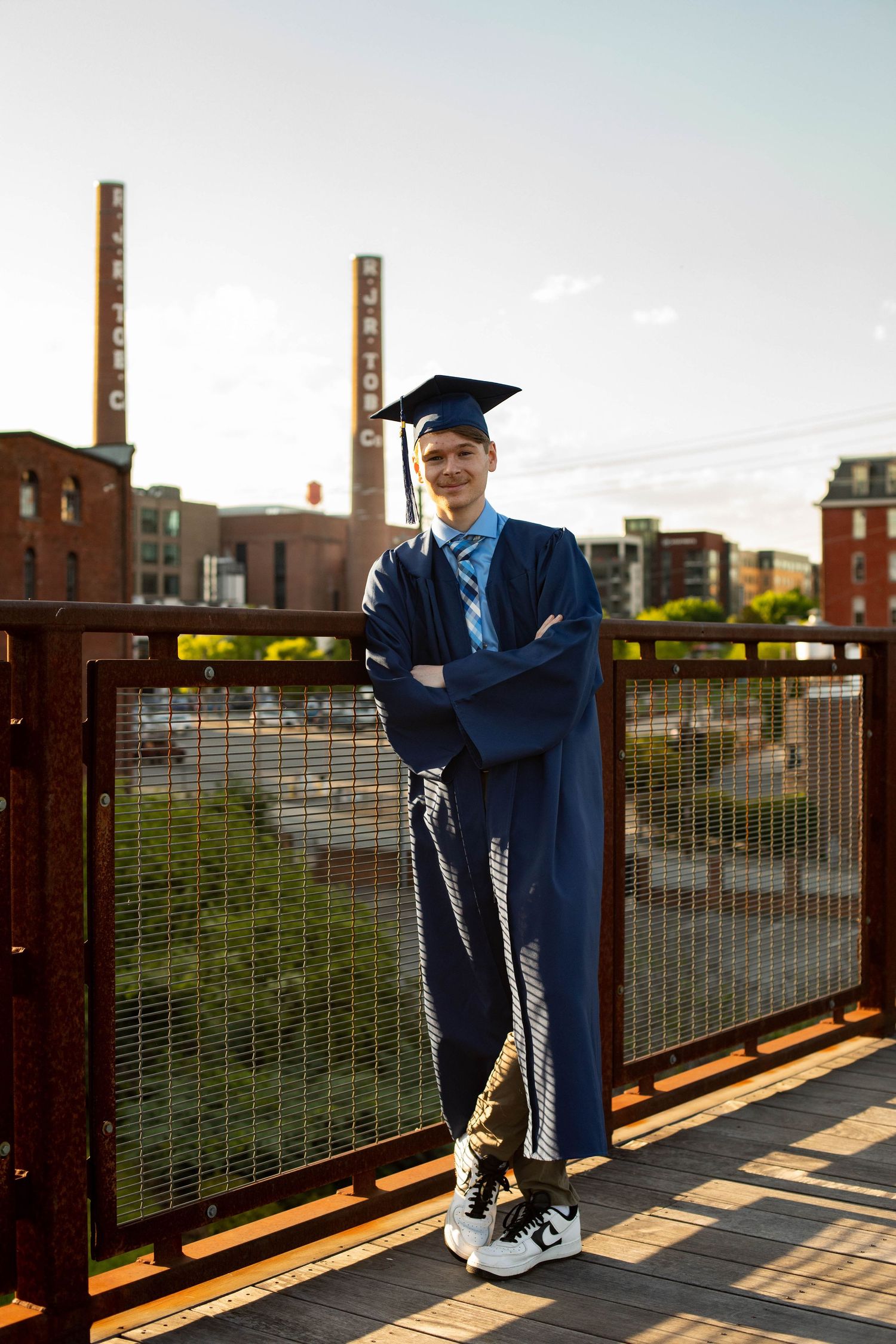 A graduate in blue cap and gown poses on an urban rooftop with industrial buildings in background.