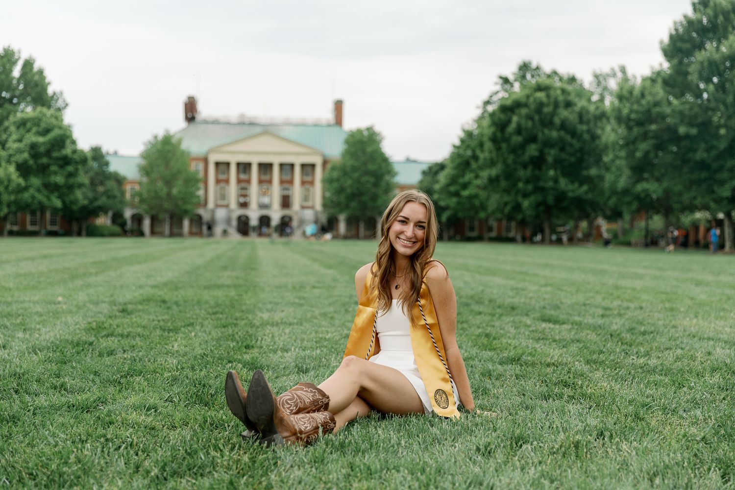 A yellow dress stands out against the lush green lawn of a grand collegiate building with columns in the background.