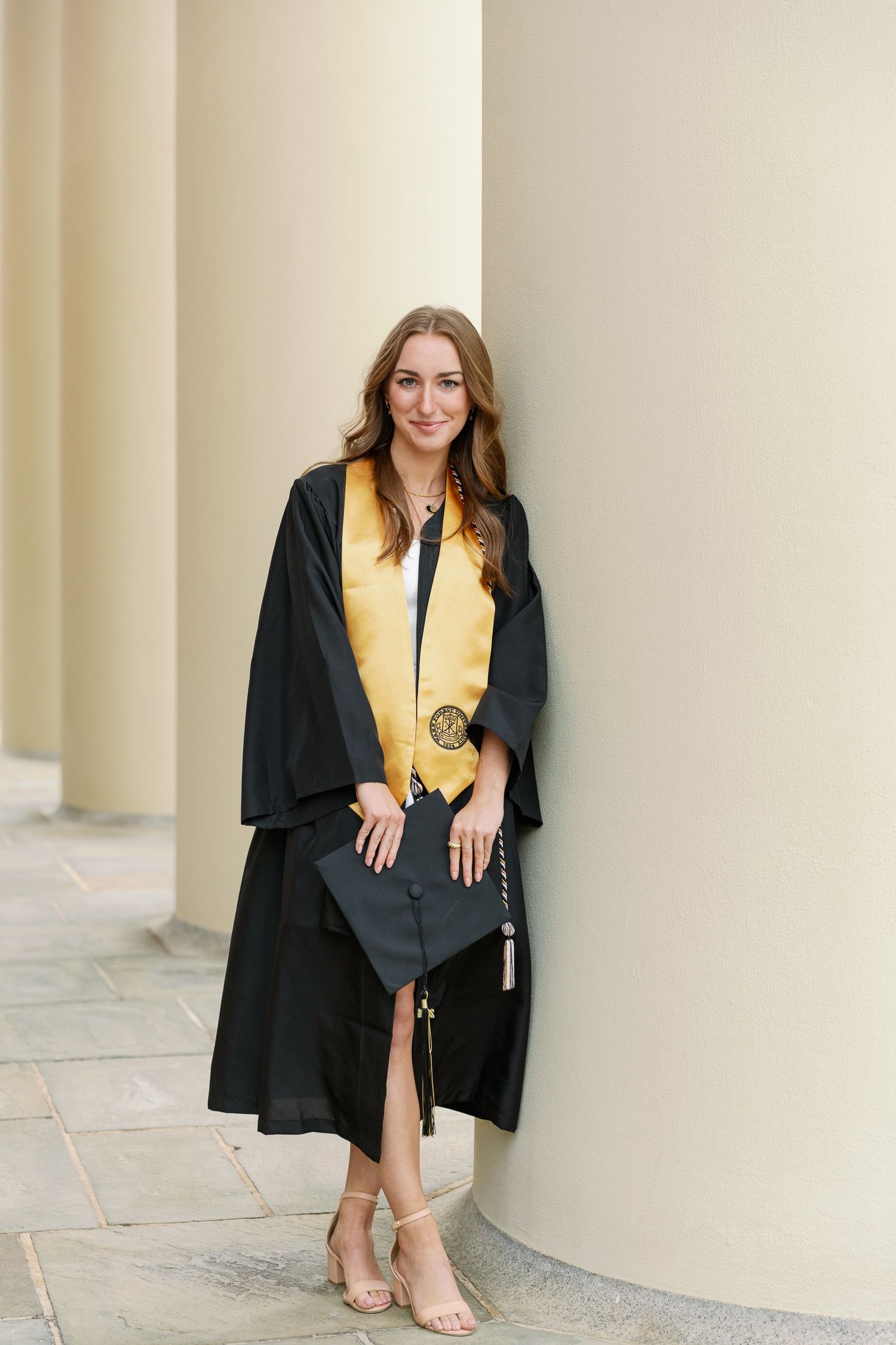 Graduate in black academic regalia and gold stole poses by cream-colored pillars.