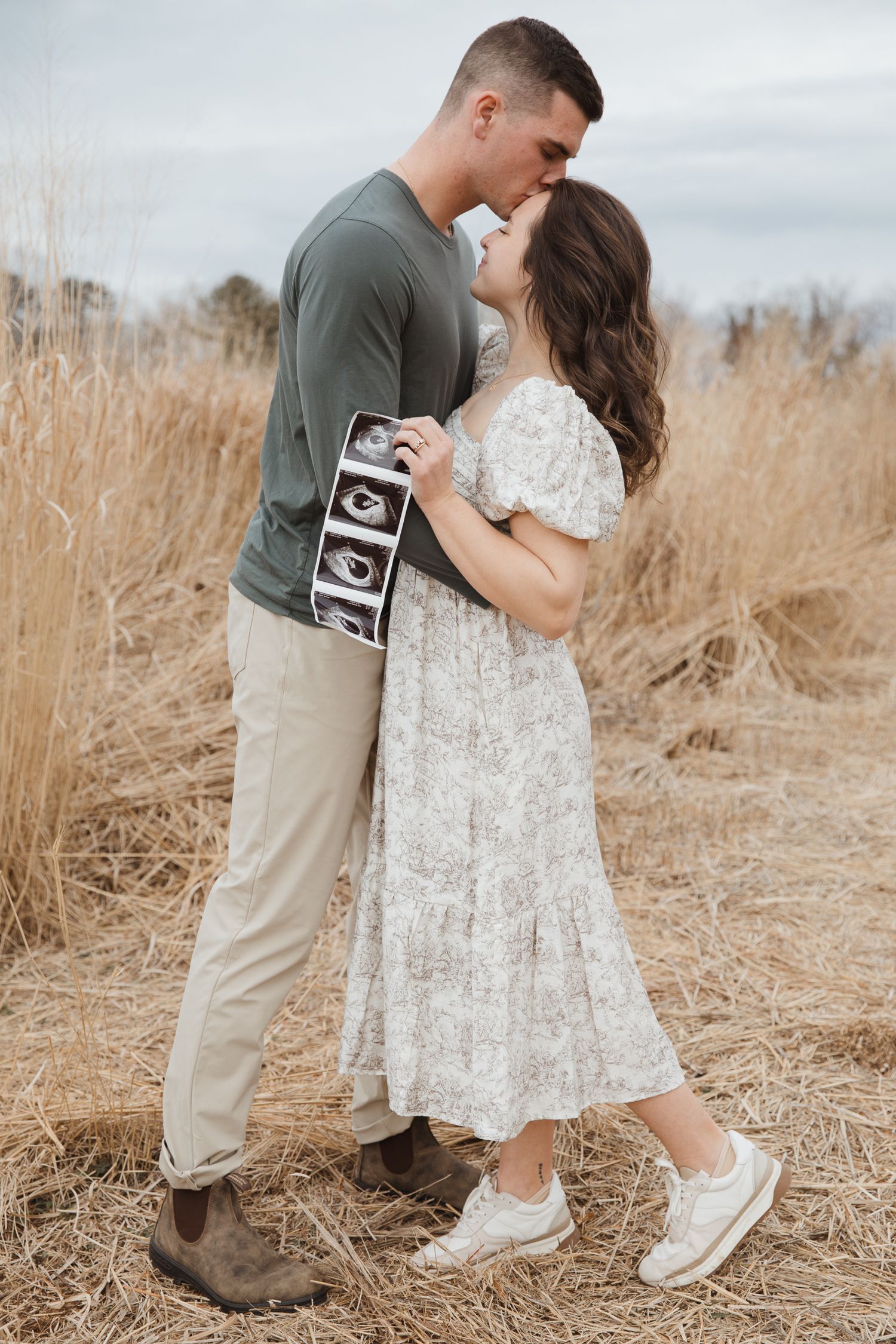 Couple kissing in a golden field, holding ultrasound photos, announcing pregnancy in a romantic outdoor setting.
