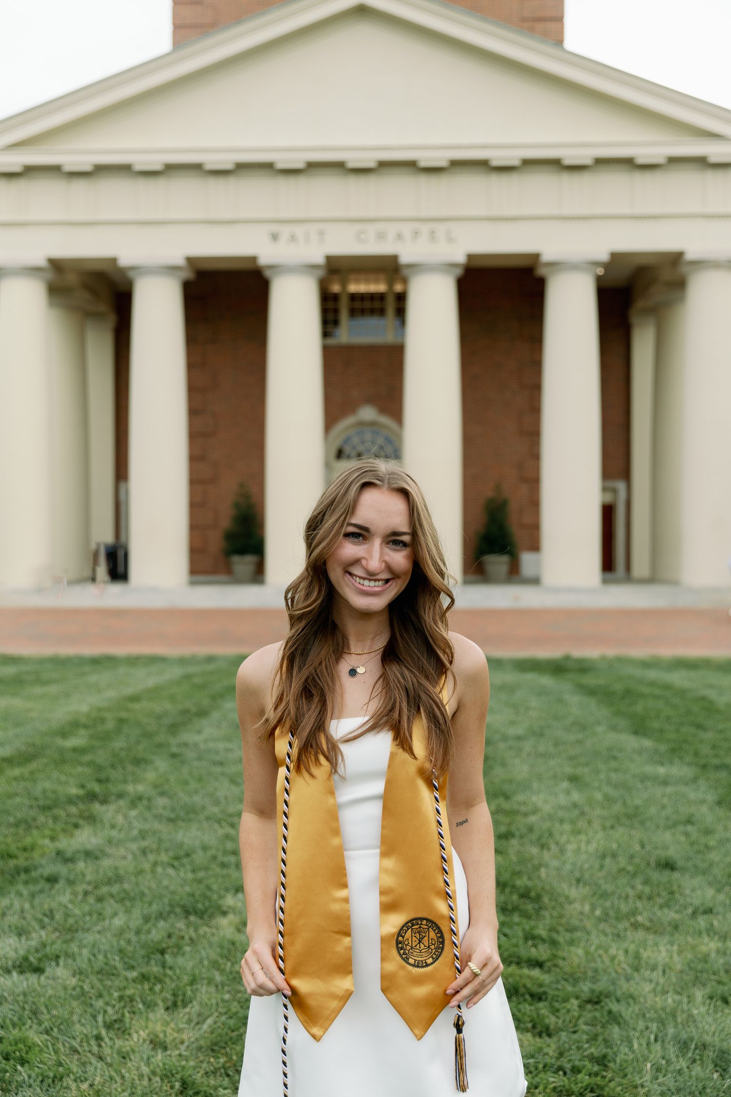 Graduate wearing white dress and gold honors stole poses in front of neoclassical building.