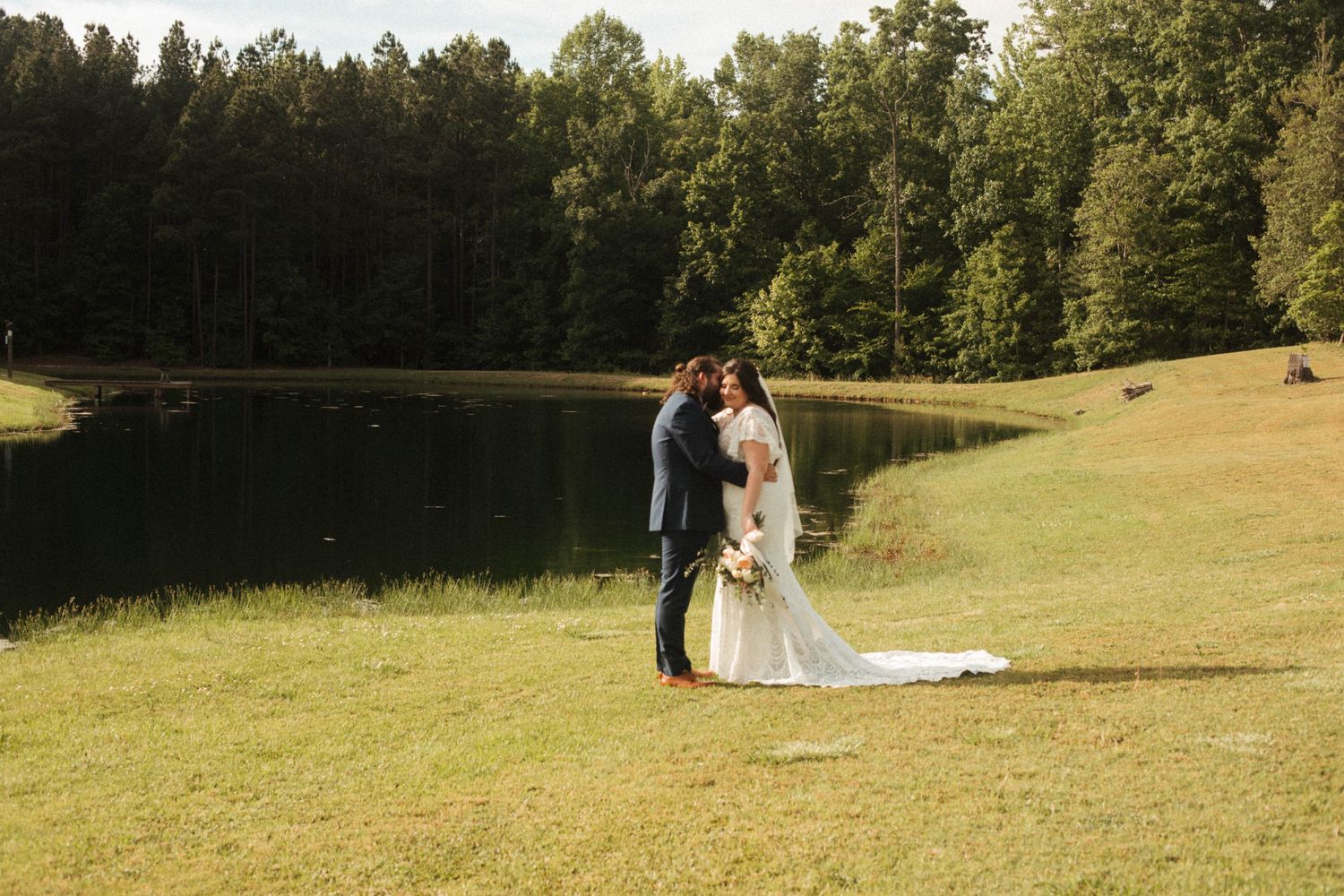 Distant couple embracing in a sunny grassy field with trees in the background.