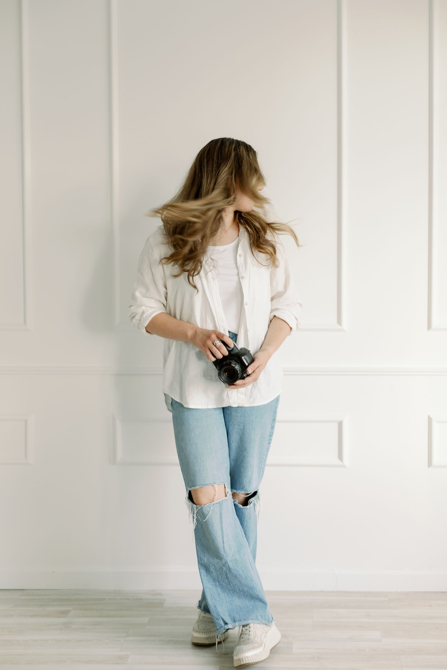 Dynamic motion shot with flowing hair, wearing distressed jeans and white layers while holding camera.