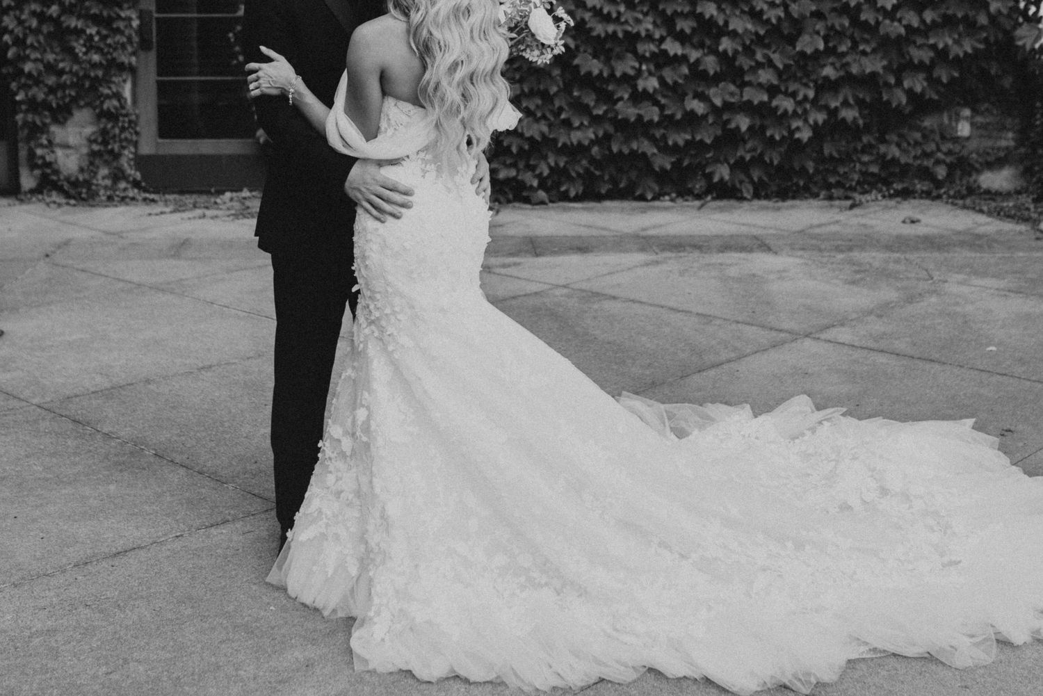 A dramatic black and white photo showcases the back of a bride's flowing lace wedding gown.