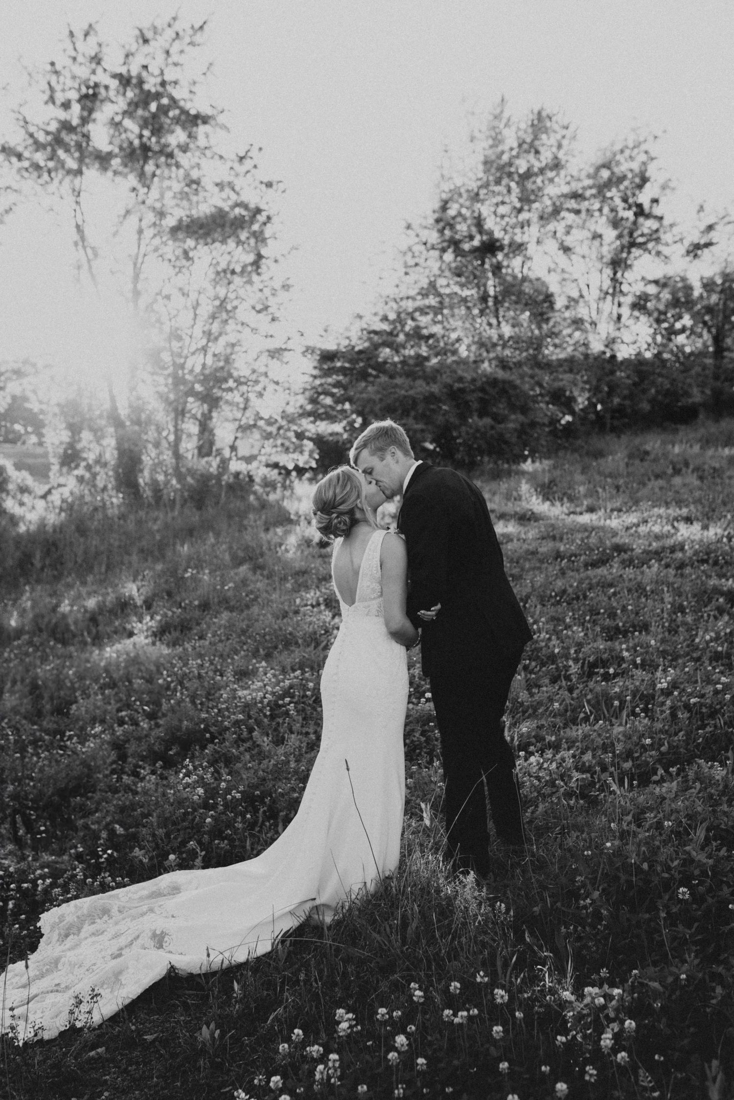 A black and white wedding photograph shows a couple embracing in a sunlit meadow with wildflowers and trees in the background.