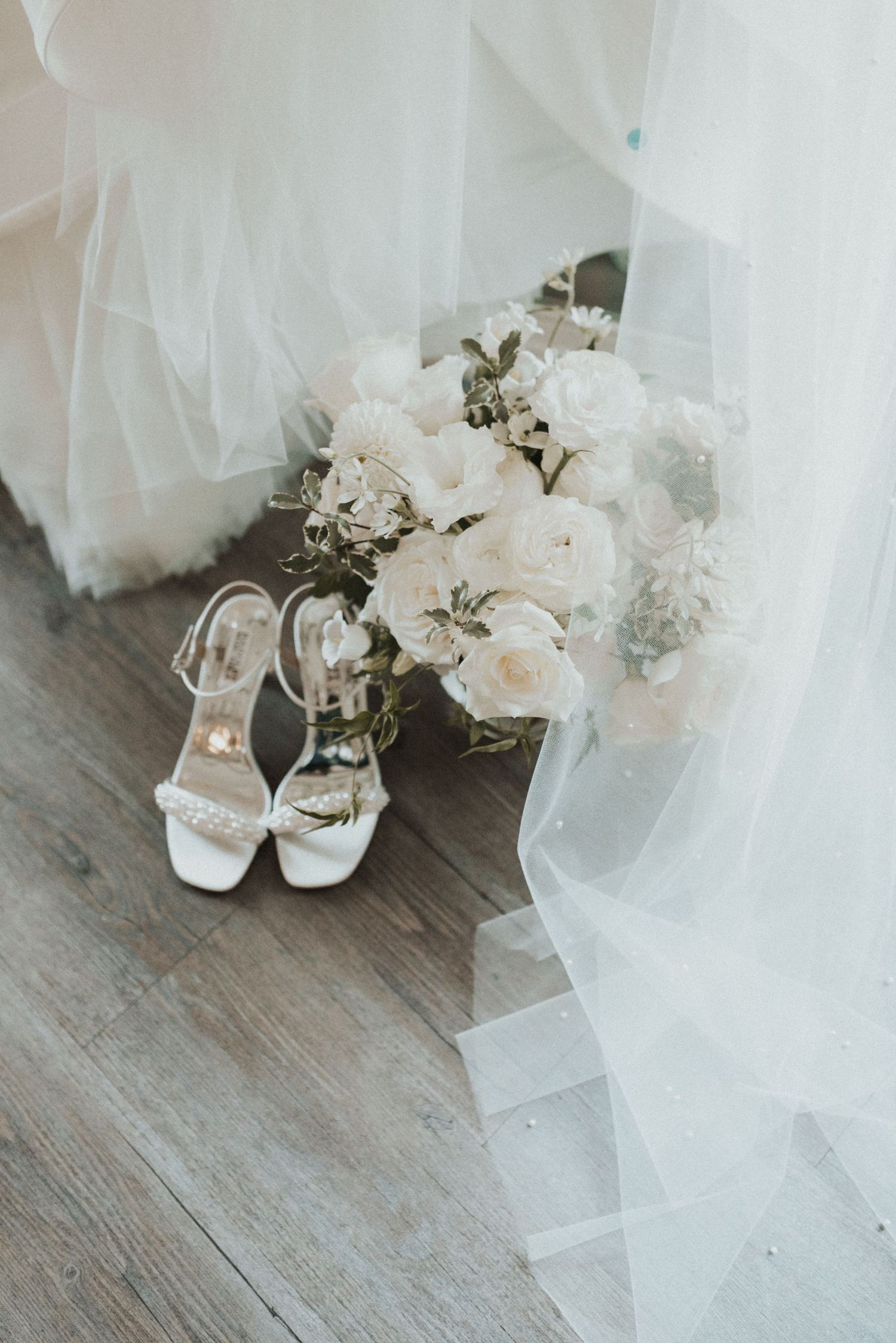 Elegant white bridal bouquet and shoes arranged with flowing tulle veil on wooden floor.