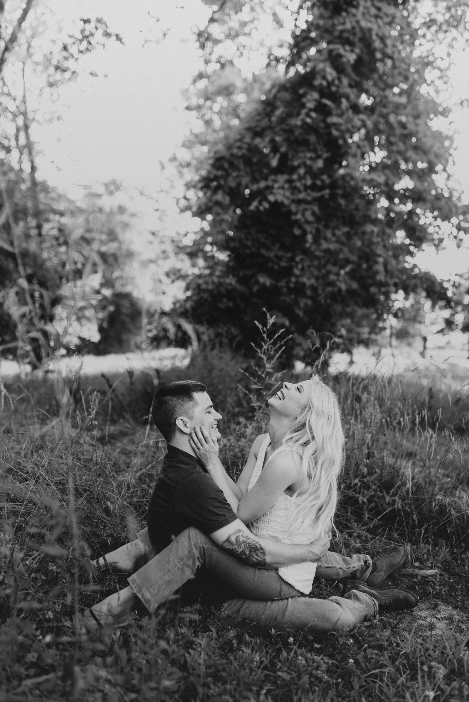 A vintage black and white photograph shows a couple sitting intimately together on grass in a pastoral outdoor setting.