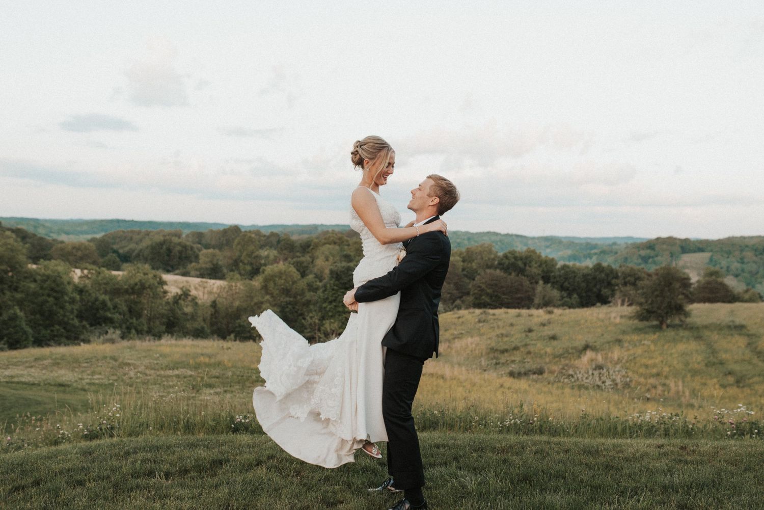 A romantic wedding photo of a groom lifting his bride in a sweeping meadow landscape.