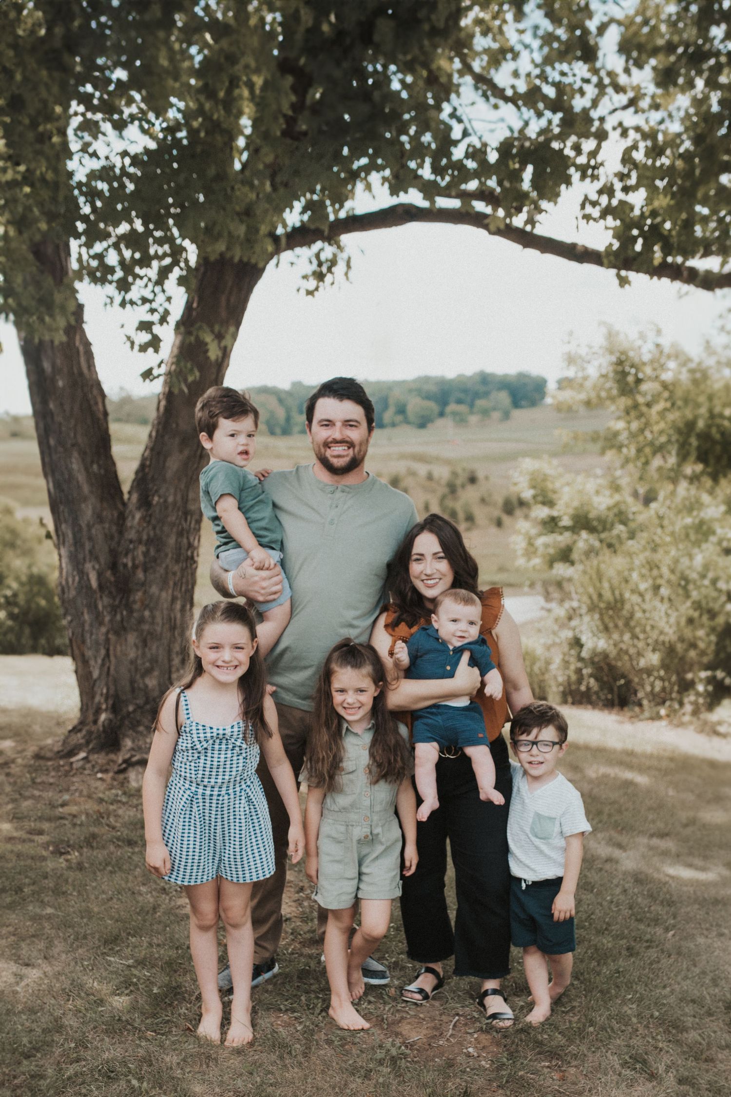 A family of seven poses together under a large tree in a pastoral setting.